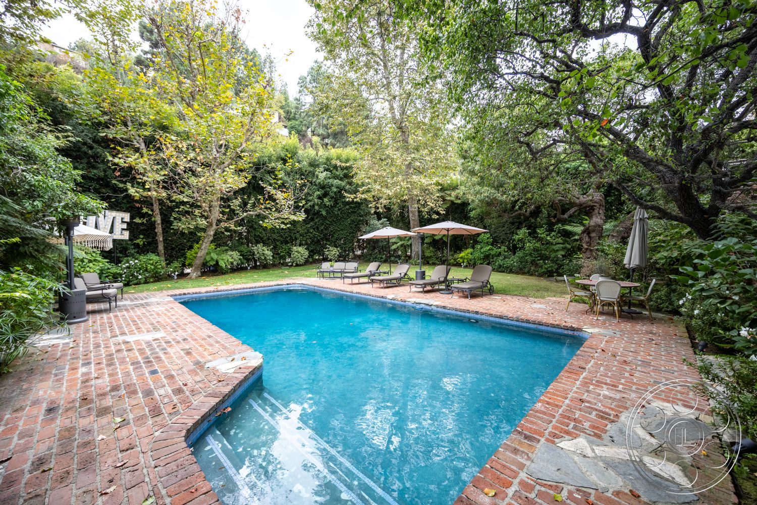 Americana 201 - rectangular swimming pool, landscaped backyard, patio seating area, brick pool deck, lush garden setting, exterior pool area, stone pool border, sun loungers with umbrellas, mature trees surrounding pool, grass lawn beside pool