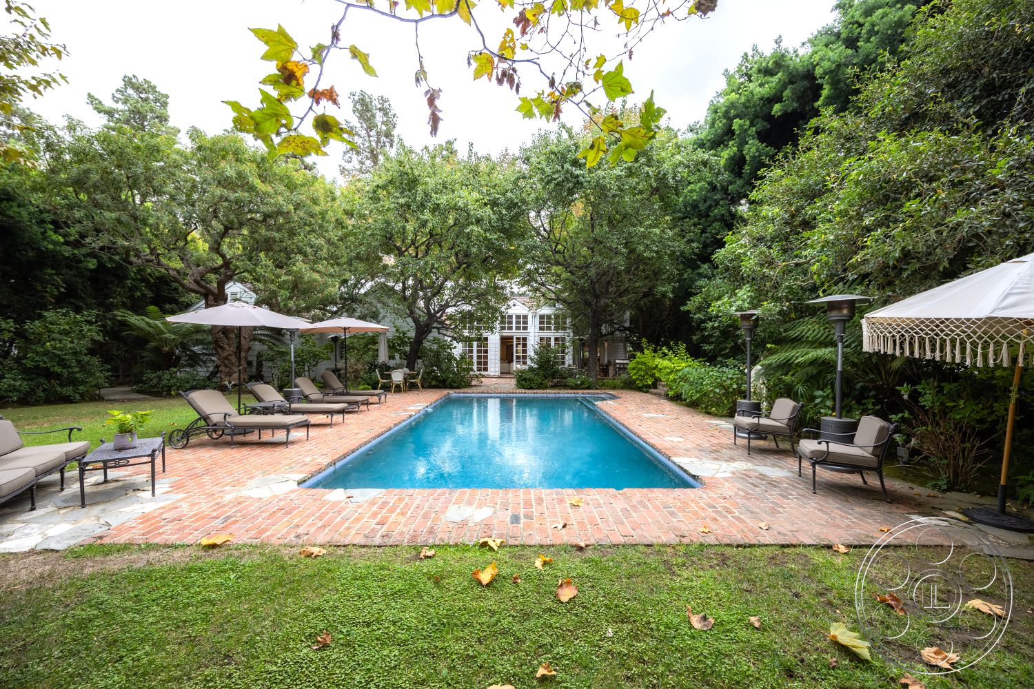 Americana 201 - outdoor seating area, rectangular swimming pool, brick pool deck, wooden fencing, landscaped garden setting, brick fireplace surround, surrounding lush greenery, stone patio flooring, exterior pool area, tropical plant landscaping, lounge chairs with umbrellas, stone pavers around pool, patio heating lamps, multi-tiered garden, poolside relaxation zone