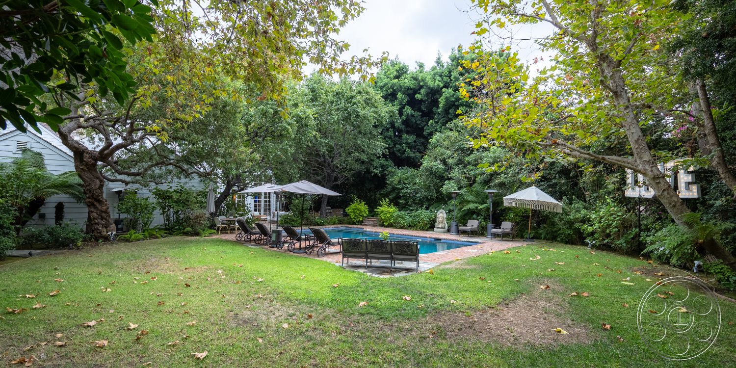 Americana 201 - exterior, landscaped garden, tranquil setting, mature trees, green lawn, shaded seating area, lounge chairs, surrounded by greenery, backyard oasis, rectangular pool, brick pool deck, poolside seating area, decorative statue, patio umbrellas, concrete pool border, lush tropical plants, pebble-stone pathway, outdoor shading structure