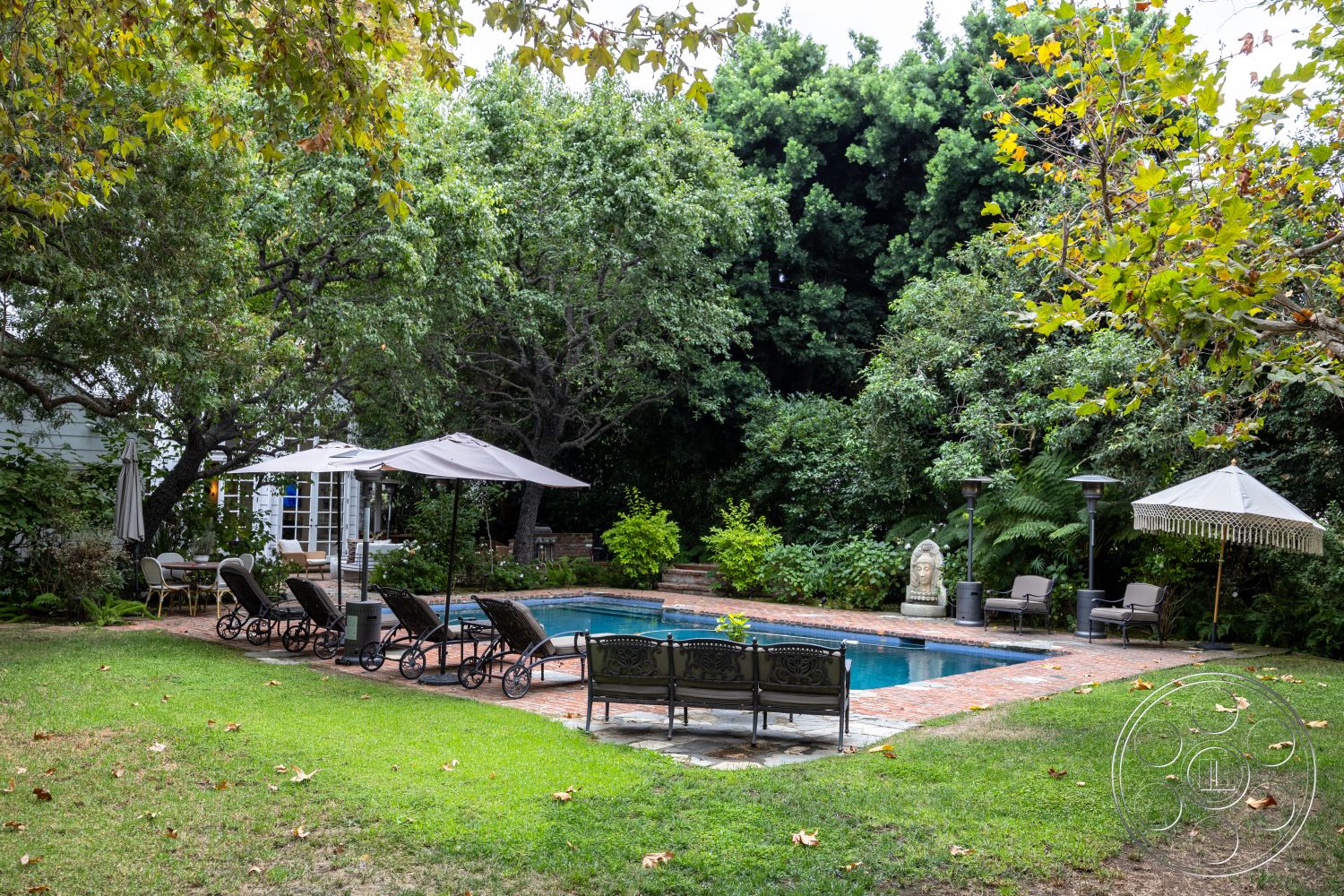 Americana 201 - rectangular swimming pool, brick pool deck, lush greenery backdrop, landscaped garden setting, concrete pool border, exterior pool area, tropical plant landscaping, lounge chairs with umbrellas, decorative stone statue accent, patio with outdoor seating, weathered wood table, relaxing outdoor retreat, sunny suburban backyard, hardscaped pathways, porch with wooden railing