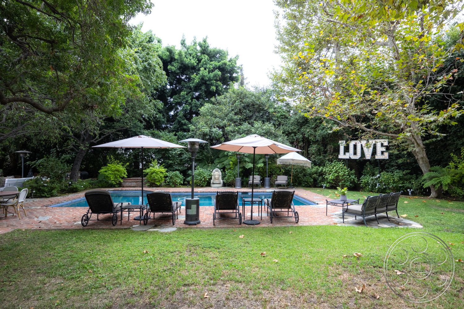 Americana 201 - exterior, rectangular swimming pool, landscaped backyard, lush garden setting, brick patio flooring, soft grass lawn, metal outdoor lounge chairs, canvas poolside umbrellas, stone garden statue, shade trees providing greenery, outdoor heating lamps, wooden garden sign, decorative pool border