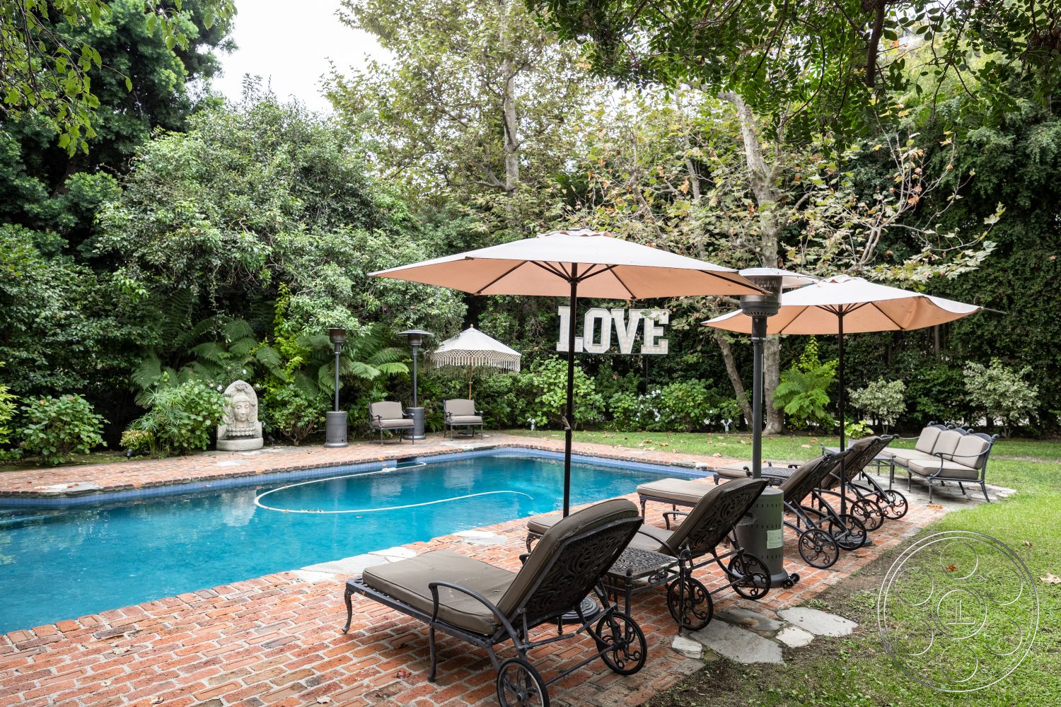 Americana 201 - rectangular swimming pool, landscaped backyard, outdoor lounge chairs, brick pool deck, poolside seating area, interior pool area, lush garden setting, natural greenery backdrop, patio heating lamps, cantilevered umbrellas, stone Buddha statue, light-gray patio stones, exposed brick planter borders, tranquil outdoor environment, decorative LOVE sign