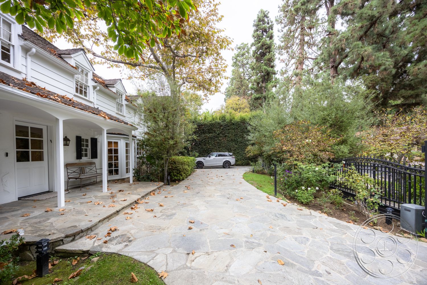 Americana 201 - outdoor lighting fixtures, decorative wrought iron fence, gabled roof with dark shingles, urban residential environment, natural stone garden edging, exterior driveway made of flagstone pavers, white painted siding, covered entryway with white columns, glass panel front door, black shutters on windows, landscape with green shrubs and flowering plants, modern automobile in driveway, leaf-strewn pathway, courtyard-style setting, tall conifer trees providing shade, curved driveway leading to garage, brick steps leading to entry