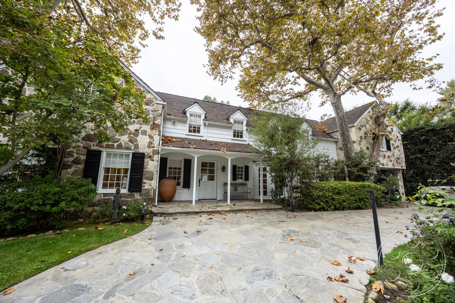 Americana 201 - transom windows, outdoor seating area, classic suburban setting, natural stone path, double-hung windows, two-story home, landscaped garden beds, white painted trim, stone exterior walls, interior courtyard view, symmetrical facade design, slate walkway, gabled roof structure, wooden porch columns, black shutter accents, front entryway porch, leafy tree canopy overhead, exposed stone details