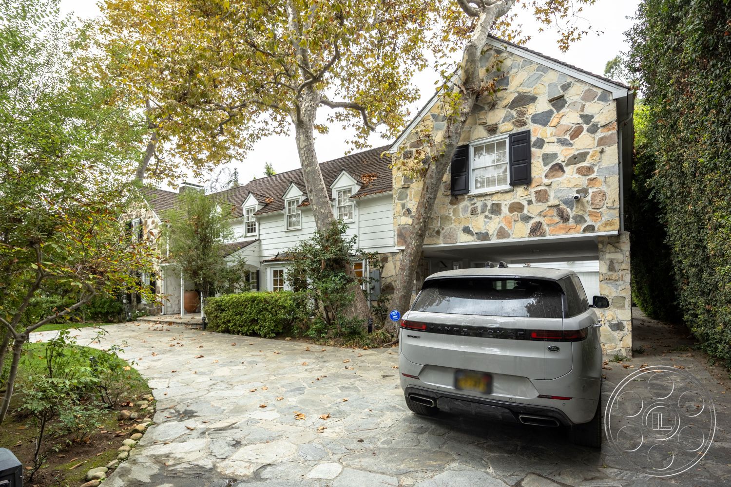 Americana 201 - wooden front door, urban residential setting, horizontal wood siding, brick wall accents, mature trees surrounding property, exterior stone veneer walls, landscaped garden with shrubbery, shingle roof house, natural stone driveway pavers, curved driveway layout, white painted wood shutters, circular stone garden bed, patio with stone flooring, two-story colonial-style home, exposed aggregate driveway, residential garage beneath house, friendly neighborhood streetscape