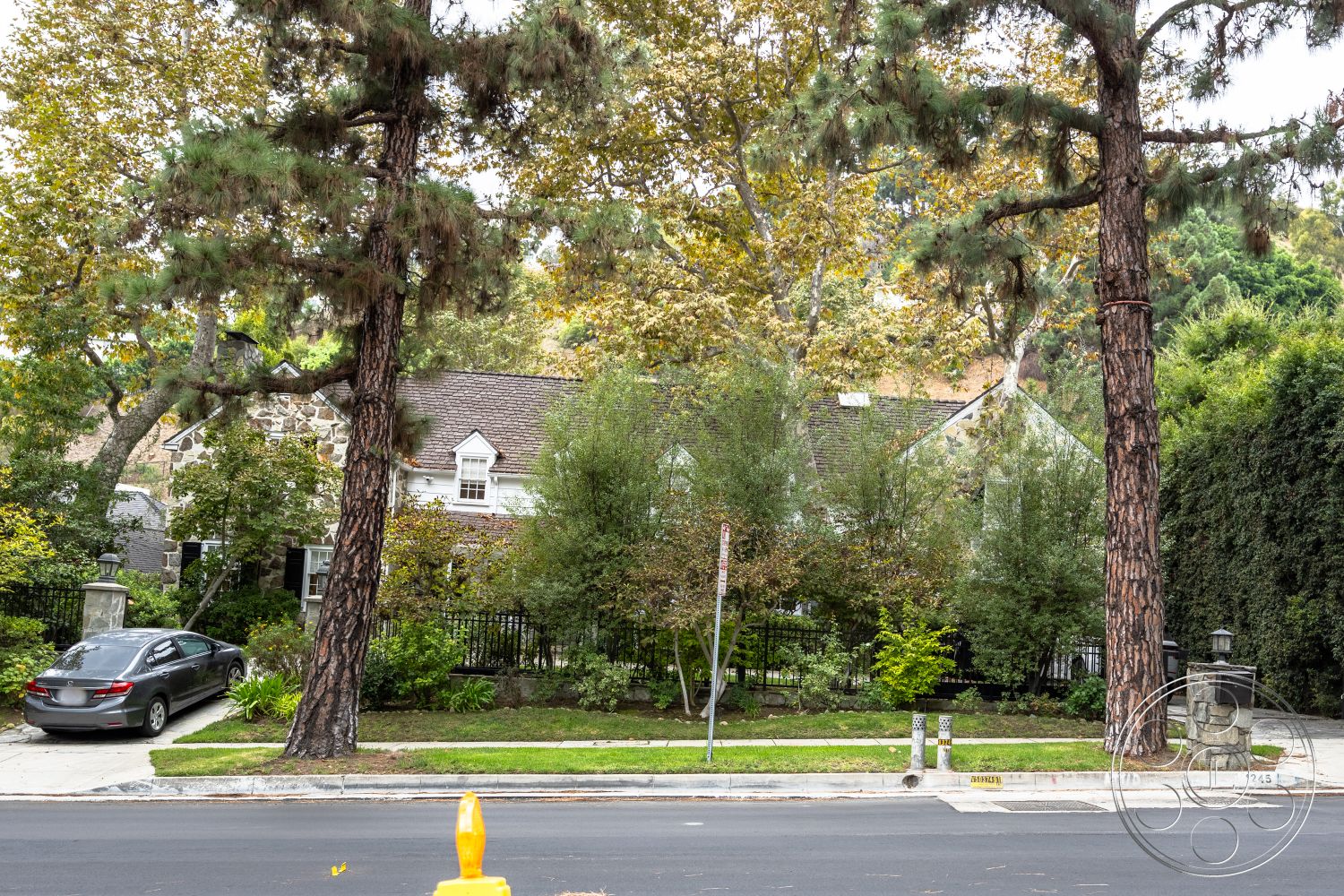 Americana 201 - exterior, fenced garden area, landscaped front yard, lush green lawn, curved driveway, natural stone pathway, decorative stone wall, city street view, hedge privacy wall, two-story home, wood shingle roof, mature pine trees, stone exterior cladding, hardwood porch flooring, urban suburban setting, decorative mailbox post, narrow window accents