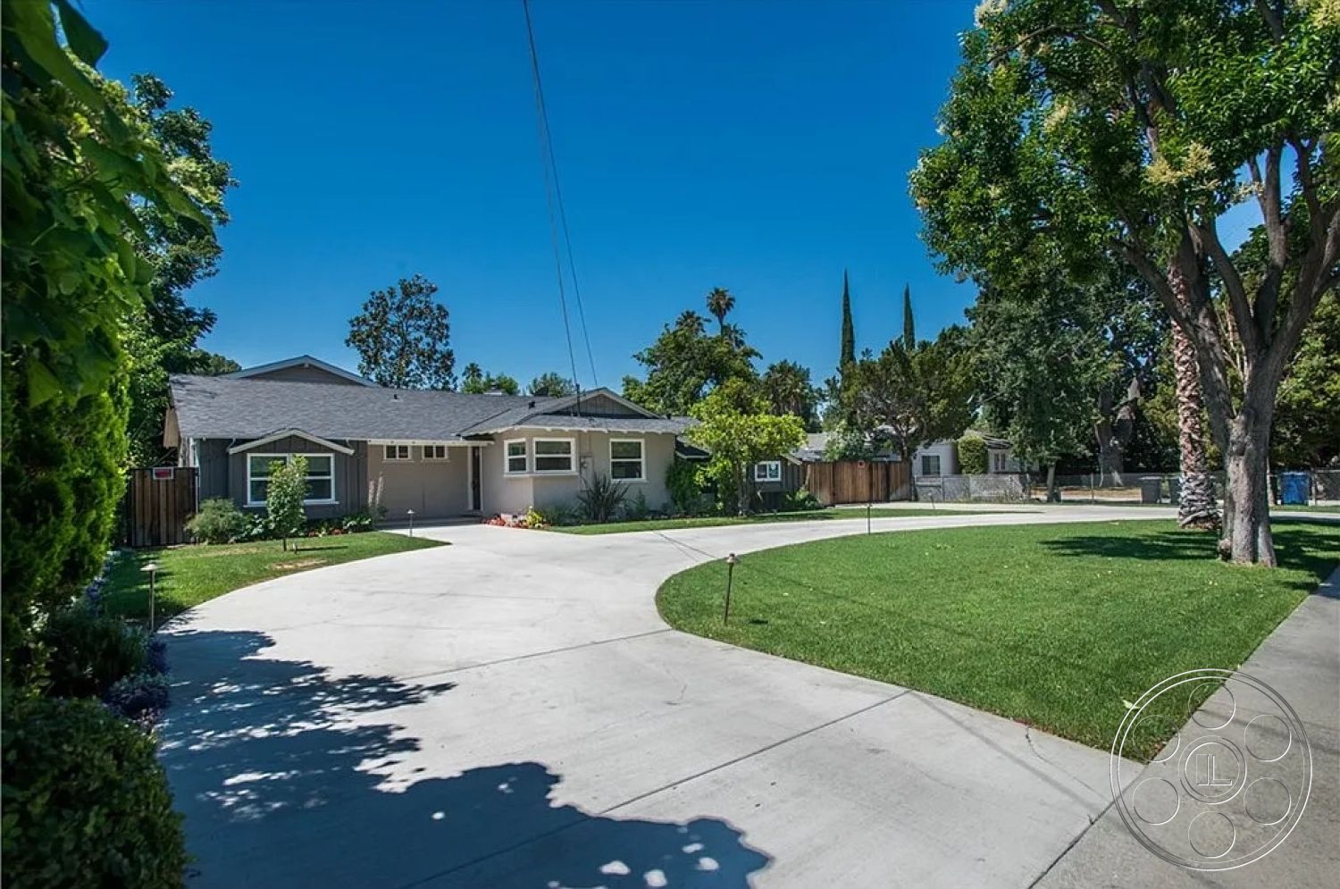 Americana 430 - tall evergreen trees, gray shingle roof, front yard garden, single-story ranch-style home, exterior wood paneling, surrounding suburban neighborhood, concrete circular driveway, grass lawn landscaping, modern window designs, dormer window details, clear blue sky setting