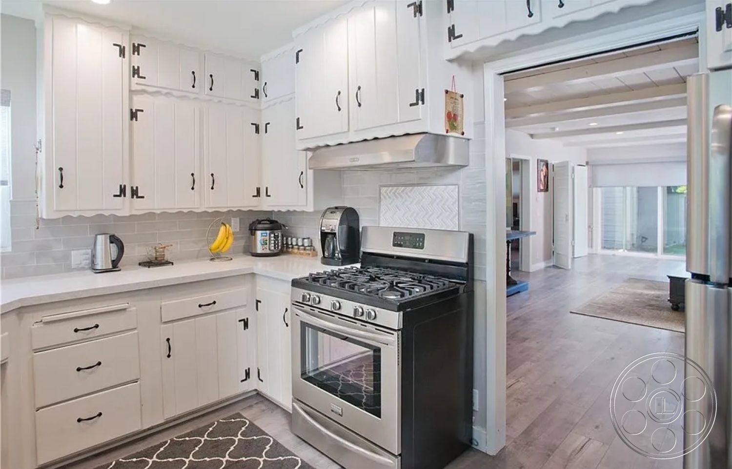 Americana 430 - light hardwood flooring, subway tile backsplash, modern farmhouse style, white quartz countertop, stainless steel kitchen appliances, functional kitchen workspace, open-concept interior layout, white shiplap cabinetry, black cabinet hardware, glass door transition to outdoor area, coffered ceiling detail, kitchen area with ample natural light, appliance garage for clutter-free aesthetics, square corner kitchen sink, decorative rug in kitchen