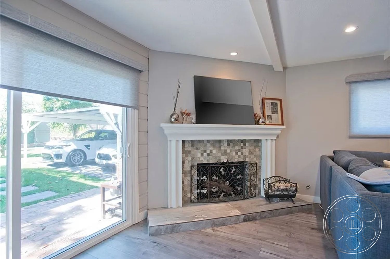 Americana 430 - minimalist decor, neutral color palette, cozy seating area, artwork on wall, open-concept layout, exposed wood ceiling beams, stone fireplace surround, light gray painted walls, hardwood living room flooring, ceramic tile fireplace hearth, large glass sliding windows, modern flat-screen television, rug under coffee table, curtains with roller shades, decorative accents on mantel