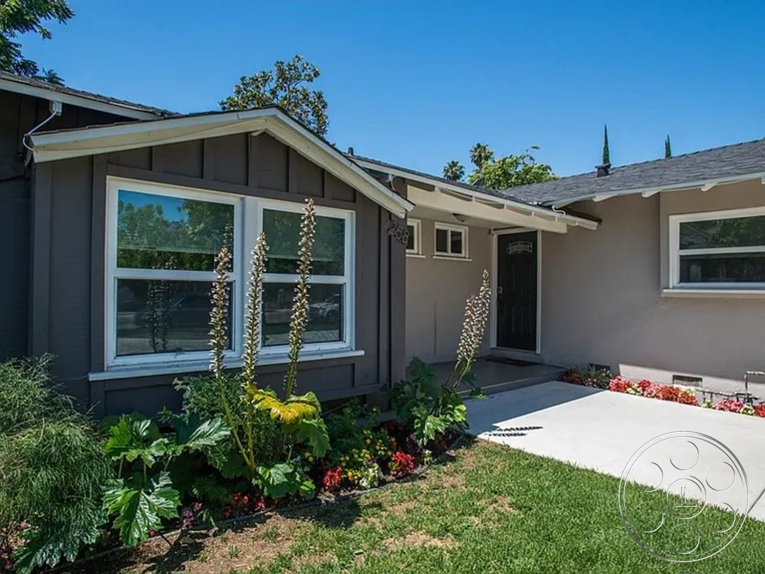 Americana 430 - exterior, suburban setting, sloped roof, green lawn, concrete walkway, wood siding, wood shutters, modern residential architecture, two-story home, overhang roof, arched front door, large double-pane windows, landscaped garden with flowers, concrete porch steps, gray wood paneling, driveway with decorative plants, arched window tops, outdoor plant beds