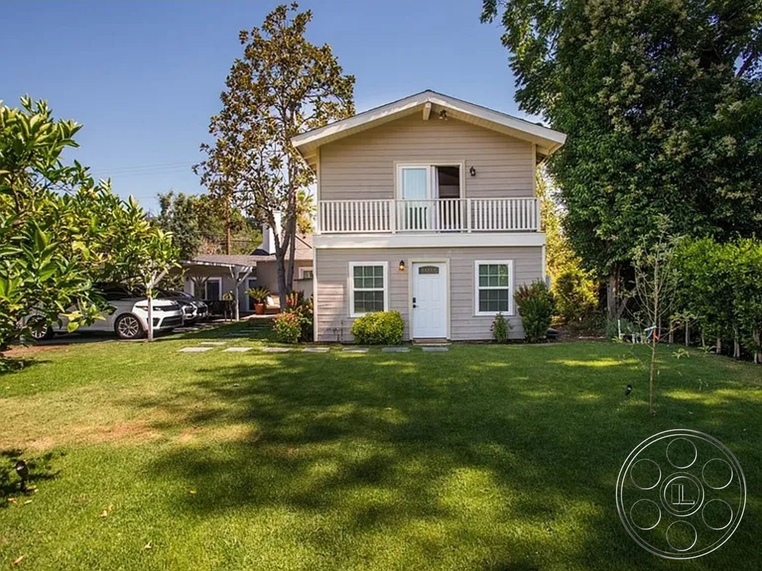 Americana 430 - exterior, surrounding trees, urban residential setting, two-story home, sloped roof design, natural light exposure, brick pathway pavers, wood siding facade, front porch balcony, white painted window trim, green lawn landscaping, decorative shrubbery, sprinkler system
