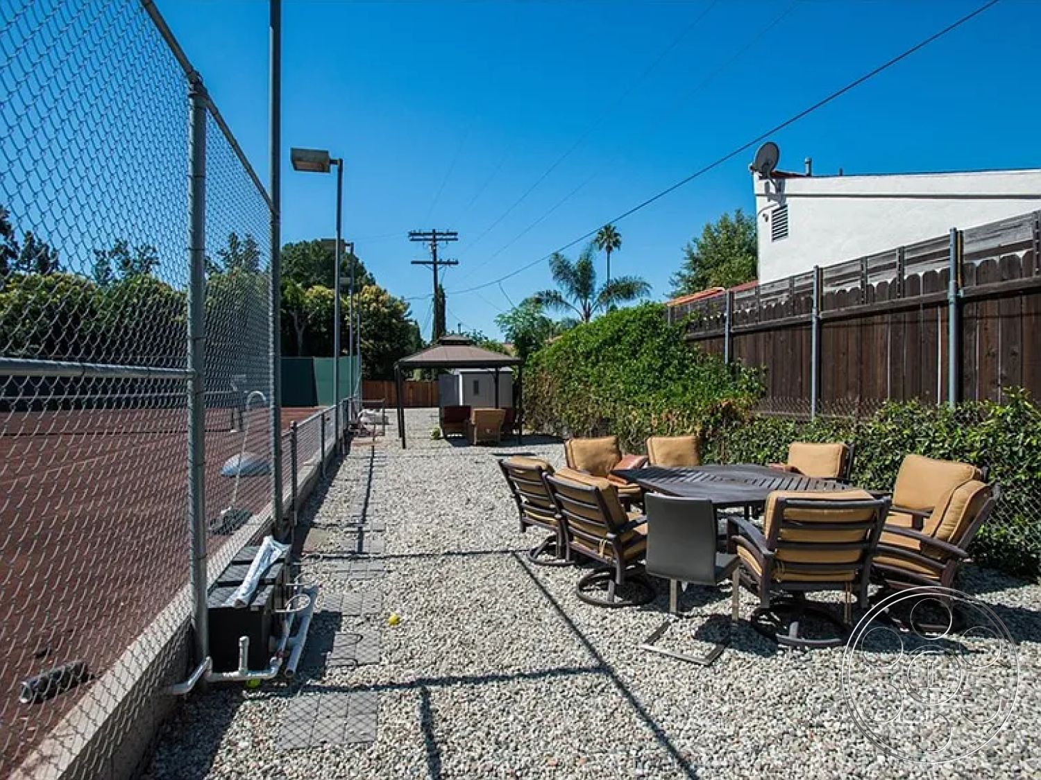 Americana 430 - paved pathway, outdoor dining set, residential backyard, chain link fence, metal frame chairs, circular dining table, surrounding palm trees, sunny outdoor setting, casual outdoor seating area, gravel patio flooring, open sky view, umbrellas for shade, concrete slab foundation, green hedge landscaping, exterior lighting fixtures, patio area adjacent to tennis court, textured gravel surface, mountainous background view