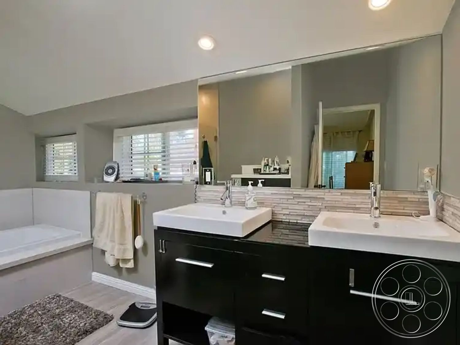 Americana 430 - textured area rug, soft gray walls, recessed ceiling lighting, interior bathroom, freestanding soaking tub, modern faucet fixtures, luxury vinyl plank flooring, white shiplap bathroom walls, modern dual sink vanity, black cabinetry with brushed nickel hardware, rectangular undermount sinks, small mosaic tile backsplash, large vanity mirror, sliding window treatments, corner bathtub with built-in platform, task lighting above mirrors, light green accent towels, shelving for bathroom essentials