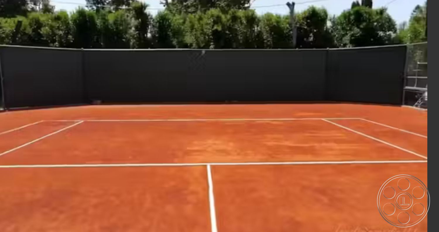 Americana 430 - sports area, outdoor recreational space, sunny outdoor setting, exterior tennis court, clay tennis court surface, red clay court with white boundary lines, surrounding privacy fencing, landscape with tall evergreen hedges, level court surface