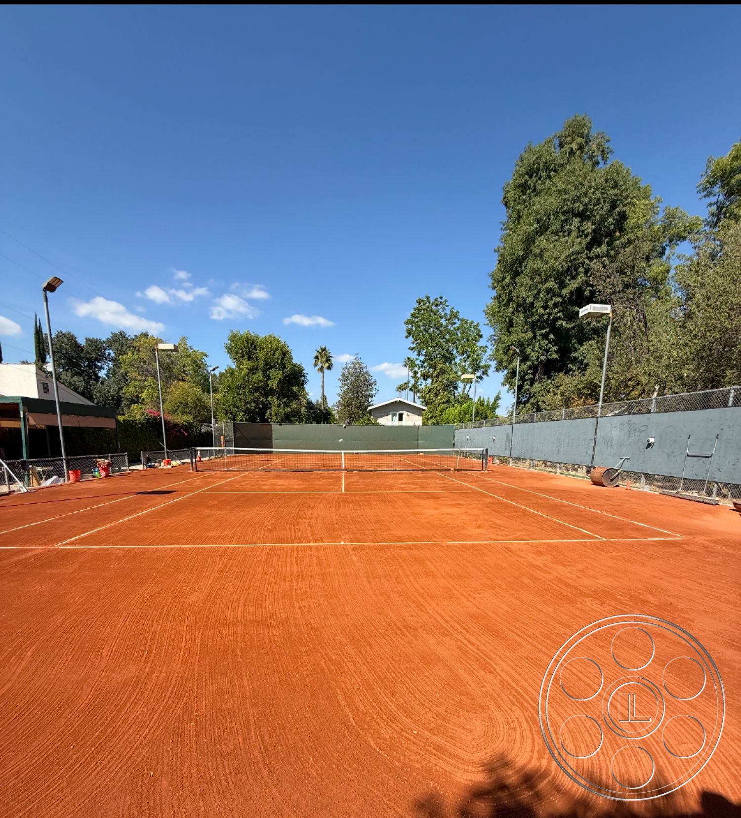 Americana 430 - blue sky, outdoor sports area, exterior lighting fixtures, residential neighborhood setting, clay tennis court surface, outdoor tennis facility, surrounding green landscaping, chain link fencing, level tennis court, planted trees in background, green fence panels, court lines marking, tennis net, natural landscape views