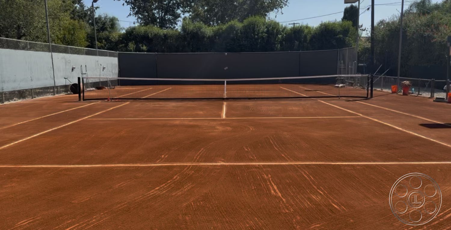 Americana 430 - surrounding trees, sunny outdoor setting, outdoor sports area, clay tennis court surface, fenced tennis court, net with tennis posts, light green boundary walls, tournament-sized tennis court, court markings in white paint, level playing surface, hard landscaping, athletic outdoor facility, residential tennis court, maintenance tools nearby