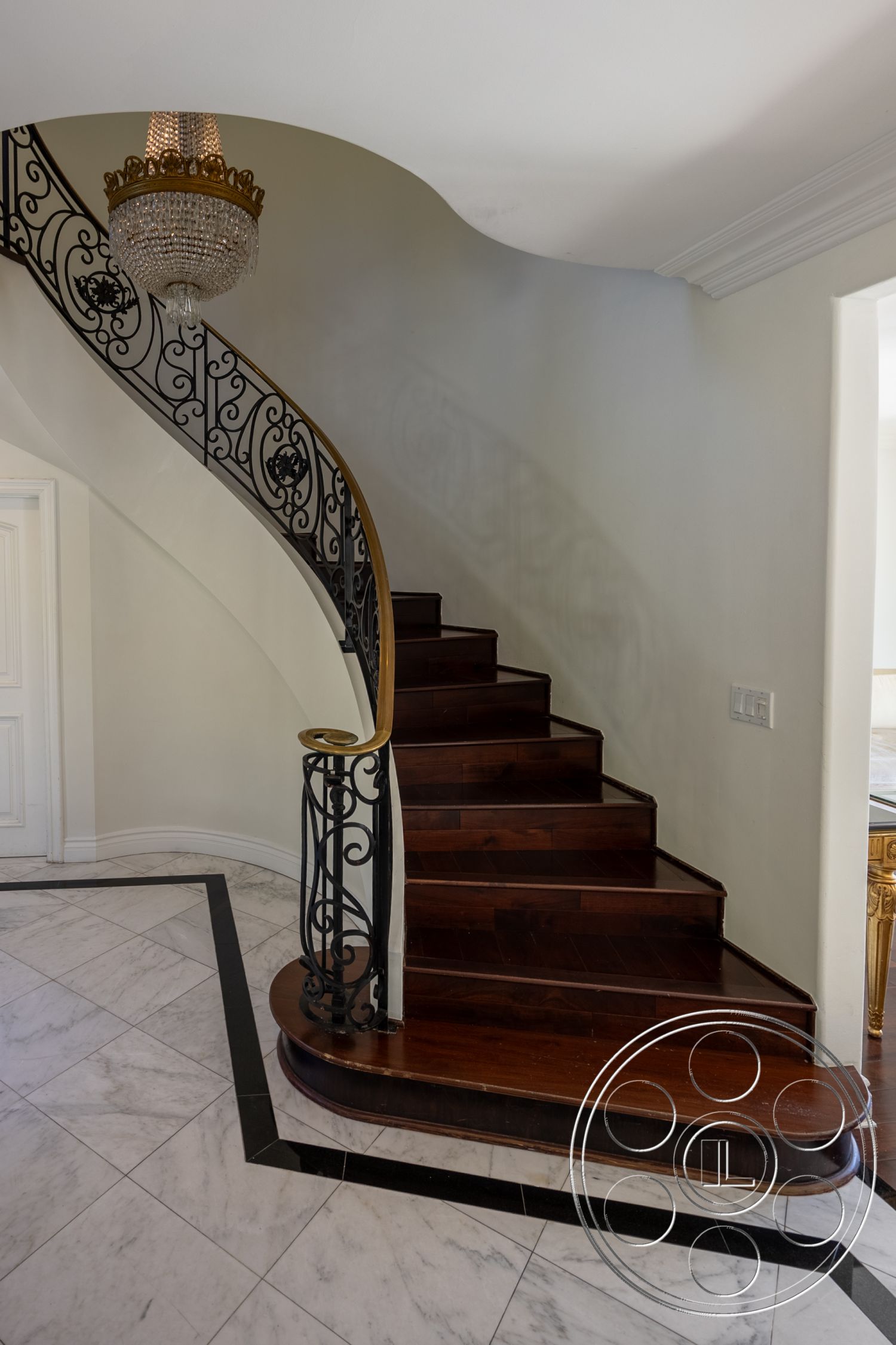 Americana 431 - cream painted walls, interior spiral staircase with hardwood steps, wrought iron railing with decorative motifs, marble floor tiles with black grout lines, curved staircase leading to upper levels, elegant chandelier with crystal details, transitional style entryway, classic crown molding details, wood risers with polished finish, open concept layout connecting to adjacent rooms
