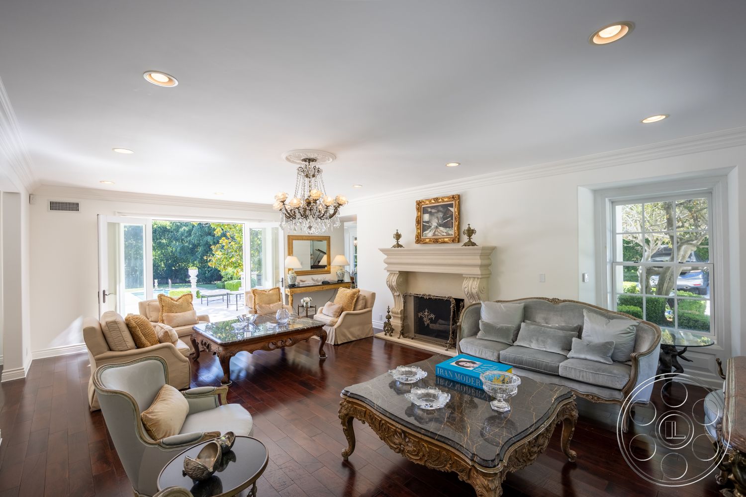 Americana 431 - glass coffee table, elegant seating arrangement, exposed wood ceiling beams, interior living space, hardwood living room floors, white painted walls, crown molding details, traditional chandelier lighting, large bay windows, ornate fireplace mantel, plush fabric upholstery, garden view from living room, gold-framed wall art