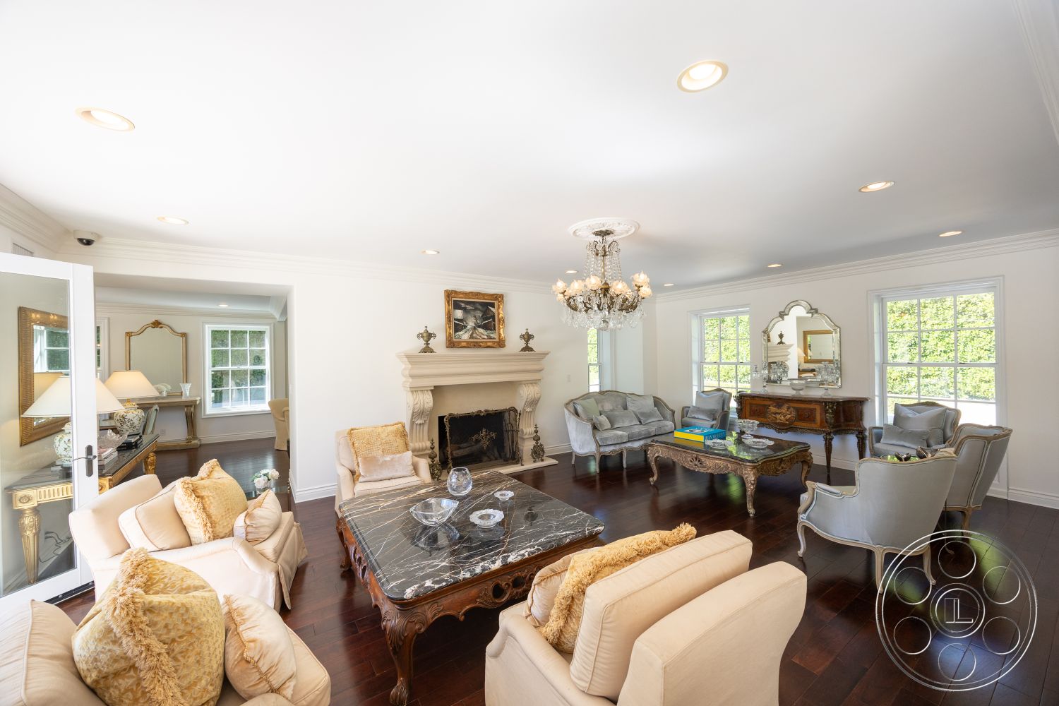 Americana 431 - marble coffee table, ornate wooden furniture, hardwood living room floors, exposed brick fireplace wall, large windows with garden view, white painted walls, coffered ceiling details, decorative wall mirror, traditional molding details, glass chandelier lighting, luxury seating area