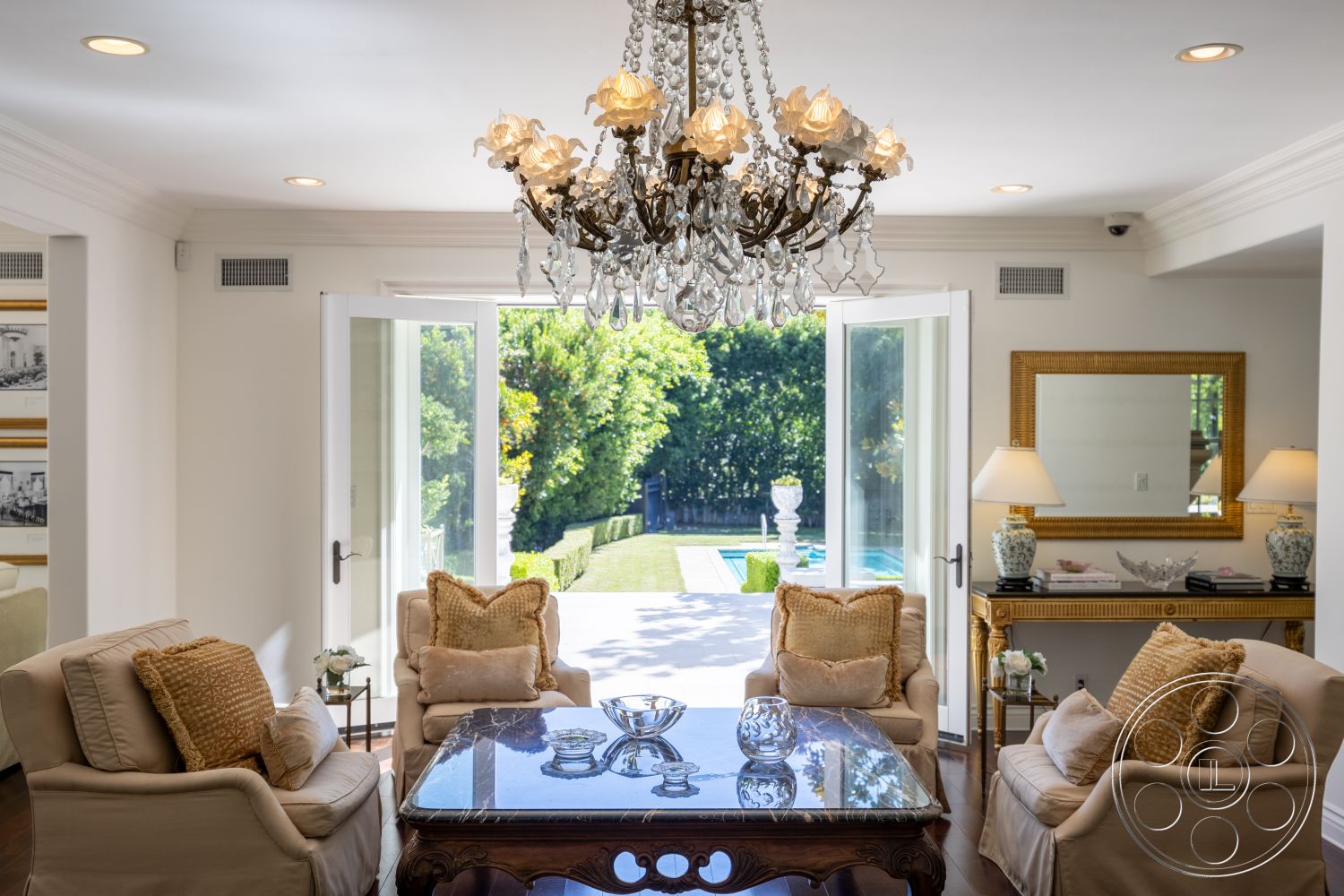 Americana 431 - marble coffee table, outdoor pool view, glass patio doors, traditional chandelier, exposed wood ceiling beams, hardwood living room floors, lush garden setting, decorative molding details, white painted walls, gold-framed mirror, plush cream sofas with accent pillows, elegant side table lamps