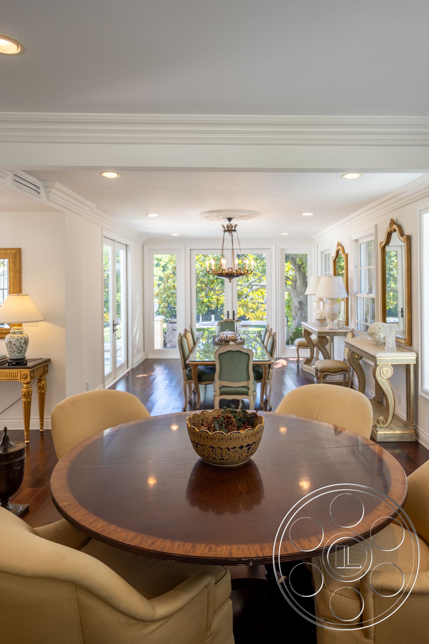 Americana 431 - indoor plant decor, interior dining area, open floor plan layout, neutral painted walls, hardwood dining room floors, large glass windows for natural light, traditional crown molding details, round walnut dining table, upholstered beige dining chairs, ornate chandelier lighting, decorative gold-framed wall mirror, elegant side console table, view of garden through windows