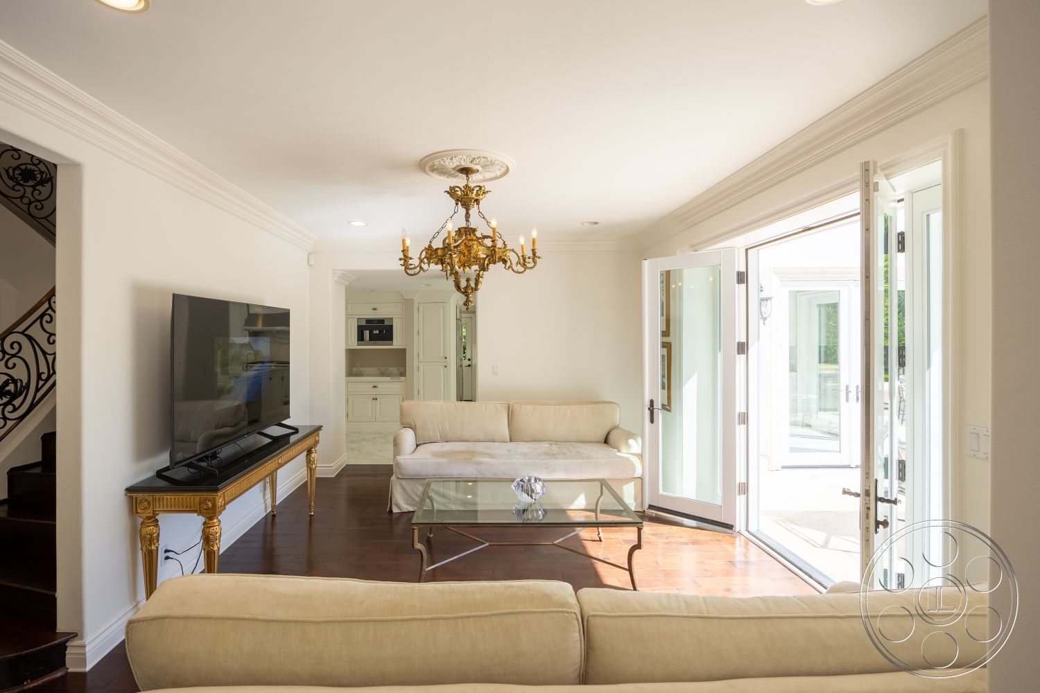 Americana 431 - hardwood flooring, glass coffee table, decorative crown molding, built-in entertainment center, comfortable lounge area, decorative wall paneling, interior living room, natural light exposure, coffered ceiling details, transitional architectural style, minimalist home decor, ornate chandelier lighting, neutral sofa upholstery, open French doors, exposed iron staircase railing