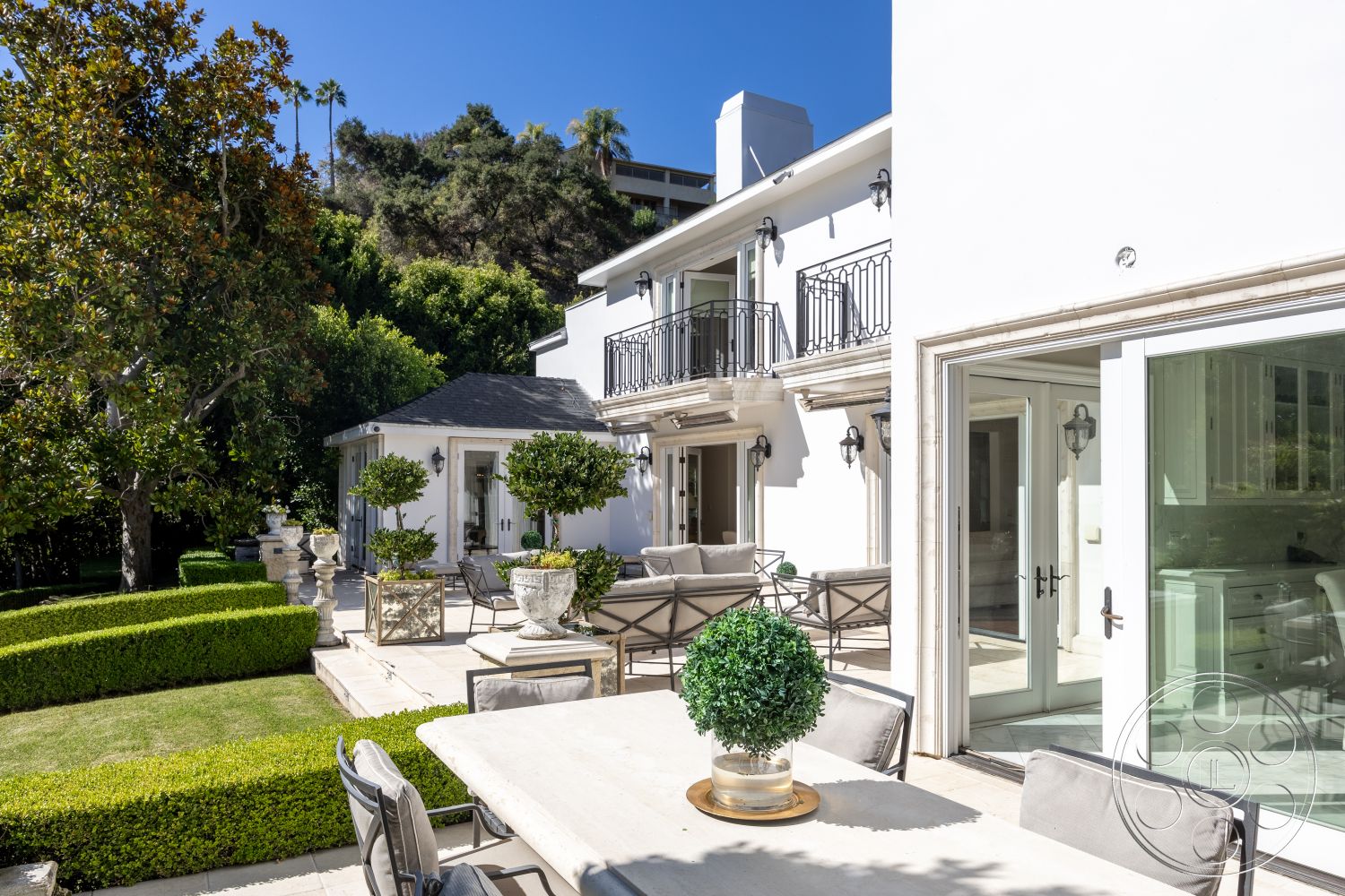 Americana 431 - exterior, large glass doors, modern outdoor decor, mountain view, trimmed hedges, balcony with wrought iron railings, metal outdoor chairs, landscaped garden area, stone patio flooring, sunlit backyard, stucco white walls, modern outdoor dining table, decorative planters with greenery, gleaming floor tiles