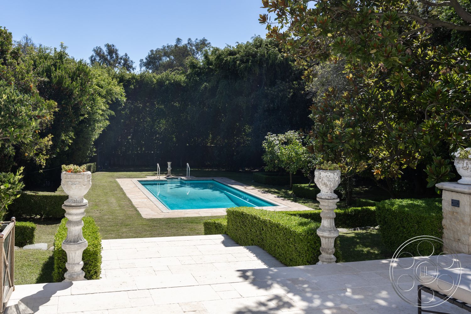 Americana 431 - rectangular swimming pool, open air space, elegant garden design, sunny outdoor setting, surrounding lush greenery, natural stone pool deck, exterior pool area, landscaped garden with hedges, stone pool border, concrete patio surface, elegant stone planters, cut grass lawn, white outdoor stone steps, classic garden elements, distant trees and foliage, tranquil backyard