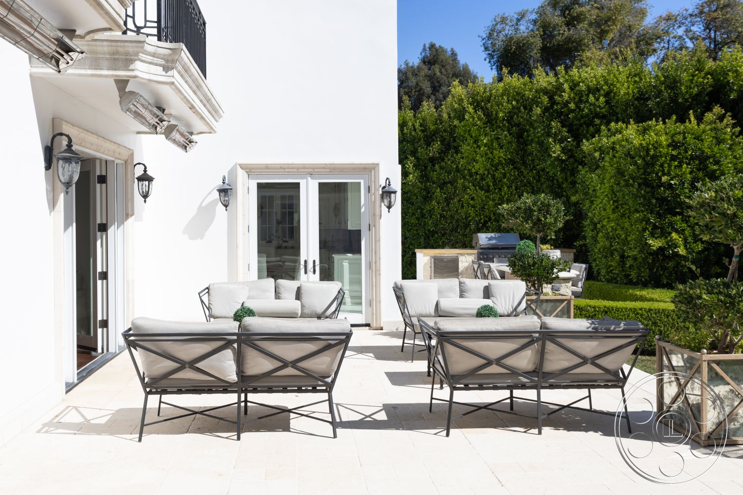 Americana 431 - white stucco walls, glass sliding doors, modern outdoor seating area, stone patio flooring, sunlit outdoor space, exterior patio space, simple geometric design, decorative planters with greenery, black wrought iron patio furniture, neutral fabric upholstery, landscaped garden surrounding, framed outdoor grill area, tropical hedge backdrop, traditional wall lanterns, minimalist exterior style, luxury home setting, inviting outdoor entertainment area