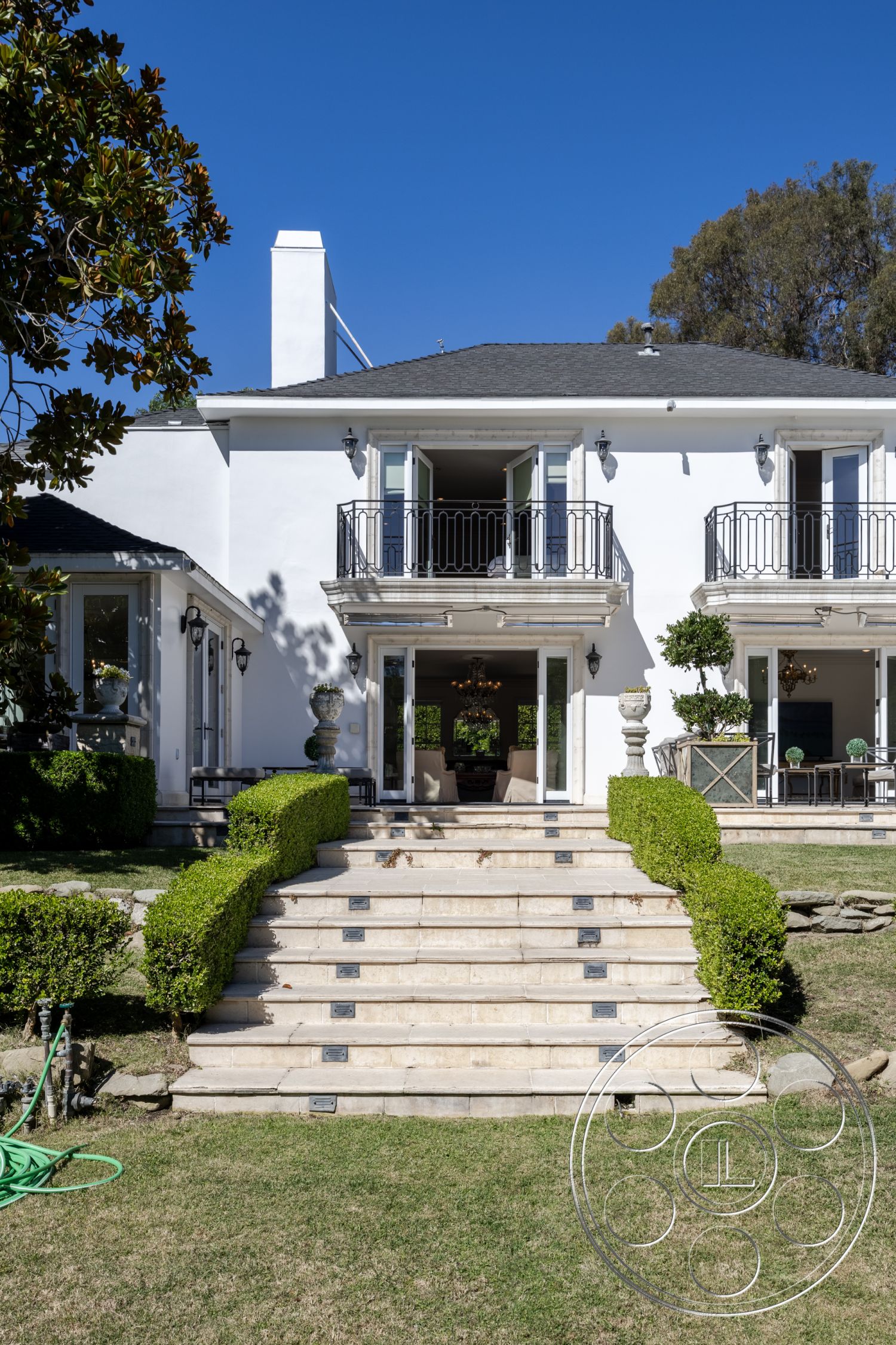 Americana 431 - exterior white stucco walls, exterior tile staircase with stone risers, exterior manicured boxwood hedges, exterior stone pathway with insets, exterior second-story balconies with wrought iron railings, exterior flat roof with overhang, exterior landscaped garden space, exterior urban home, exterior formal entryway, exterior ornamental garden urns, exterior large glass doors leading into living space, exterior balanced and symmetrical facade, exterior decorative chimney structure