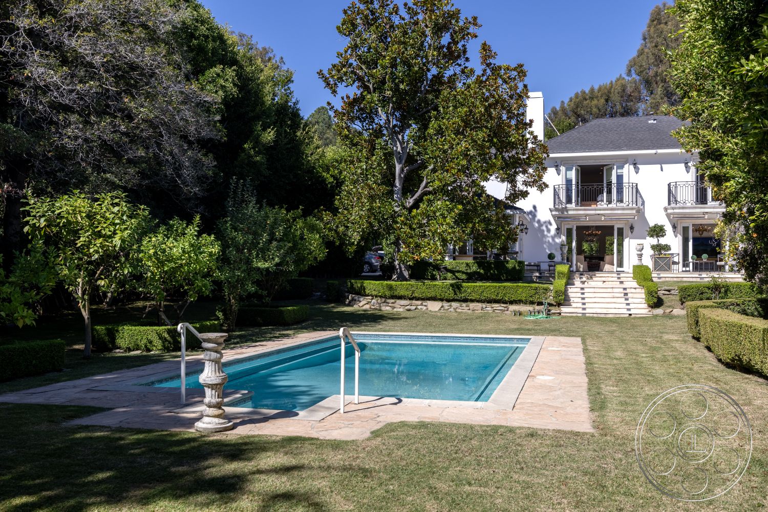 Americana 431 - exterior, formal garden layout, rectangular pool, stone pool deck, lush greenery setting, mature trees providing shade, natural stone wall accents, concrete patio border, landscaped garden with manicured hedges, three-story white stucco house, balcony with black iron railings, stairs leading to main entrance, paved poolside area, large windows with plantation shutters, symmetrical architectural design, well-maintained grass lawn, classic outdoor lighting fixtures, large backyard space