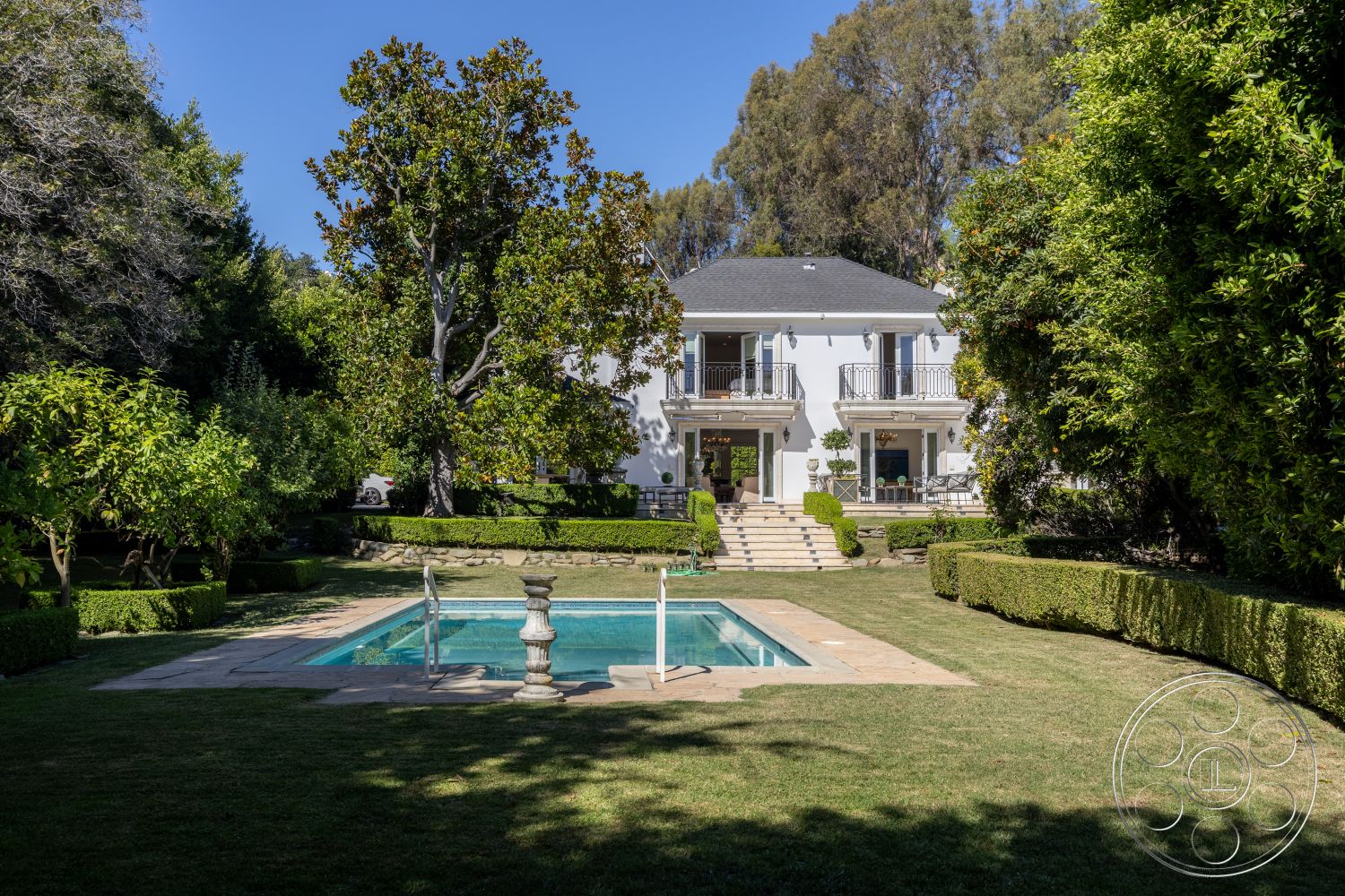 Americana 431 - exterior, stone walkway, manicured lawn, surrounded by greenery, garden setting, traditional architectural style, rectangular pool, balcony with wrought iron railings, hedge landscaping, stone pool deck, dark roof shingles, two-story home, white painted walls, concrete pool steps, patio area with outdoor furniture, steps leading to the house, urban suburban blend, outdoor space for entertaining, large windows with glass doors, tall trees providing shade