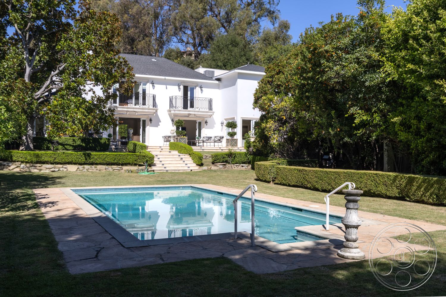 Americana 431 - rectangular swimming pool, balcony with wrought iron railings, two-story home, white painted exterior walls, exterior concrete pool border, gabled roof design, natural stone patio flooring, lush green surroundings, sunny residential setting, landscaped garden areas, brick steps leading to patio, decorative stone wall around garden, landscape hedges, outdoor furniture seating area