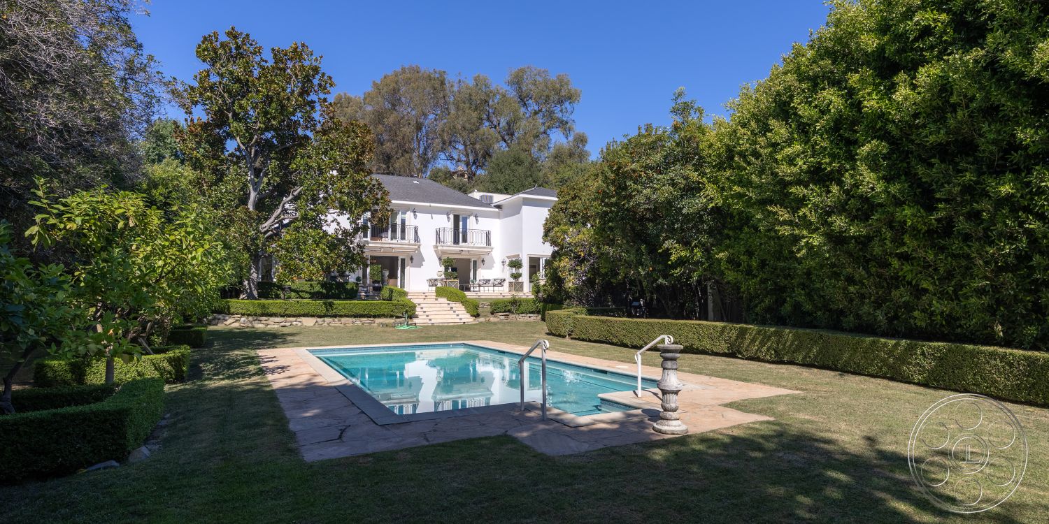 Americana 431 - exterior, manicured lawn, surrounded by greenery, white exterior walls, circular pathway, traditional architectural style, rectangular pool, poolside lounging area, sunny outdoor setting, two-story home, landscaped garden area, stone patio flooring, concrete pool border, low-maintenance hedges, balustrade steps