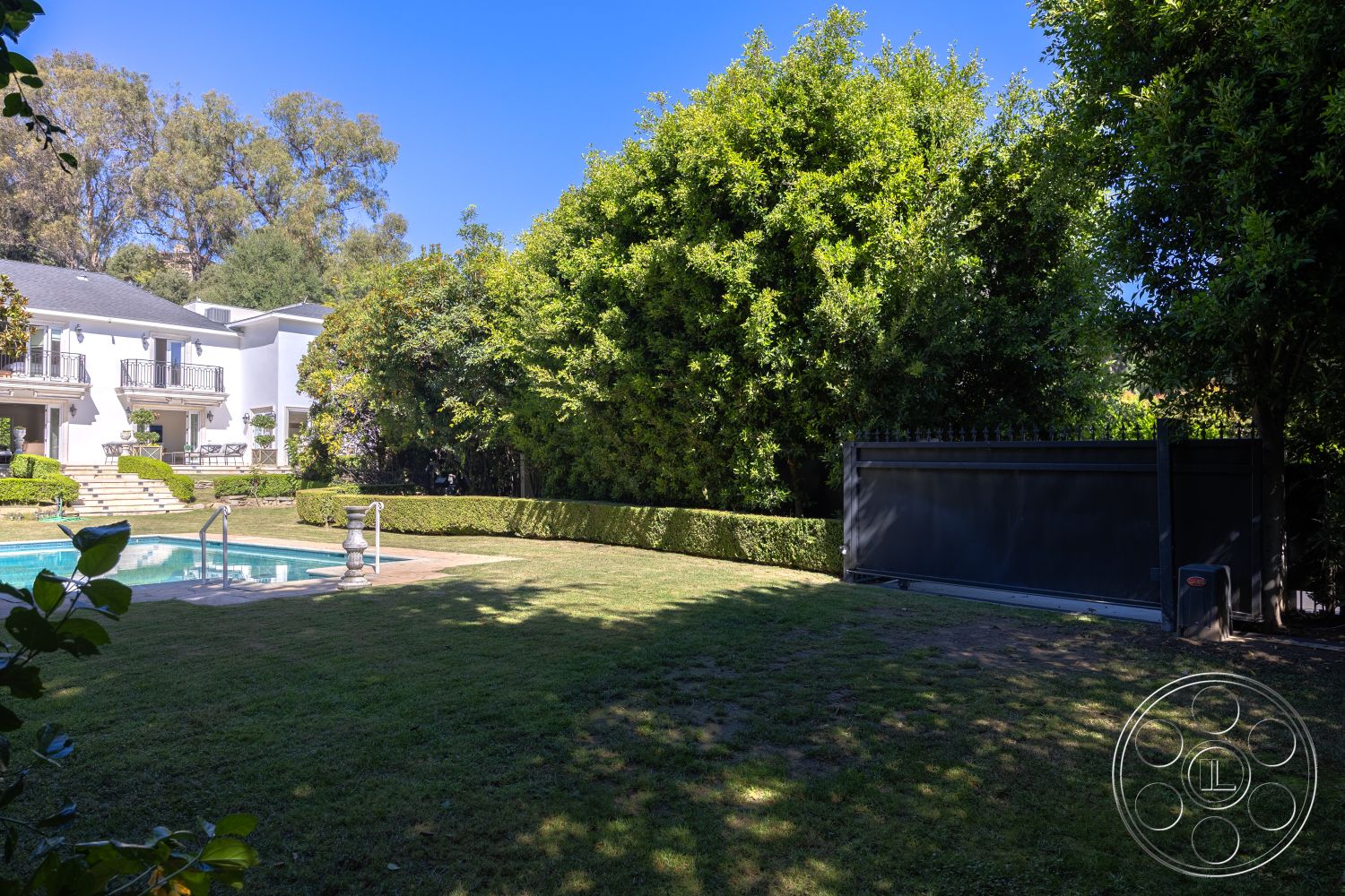 Americana 431 - exterior, white stucco walls, outdoor seating area, lush green lawn, manicured hedges, urban residential setting, balcony with wrought iron railings, stone pool deck, privacy gate, two-story home, concrete pool border, city skyline view in distance, surrounding landscaped garden, tall trees providing shade, inground rectangular pool, paved stone steps leading to the pool