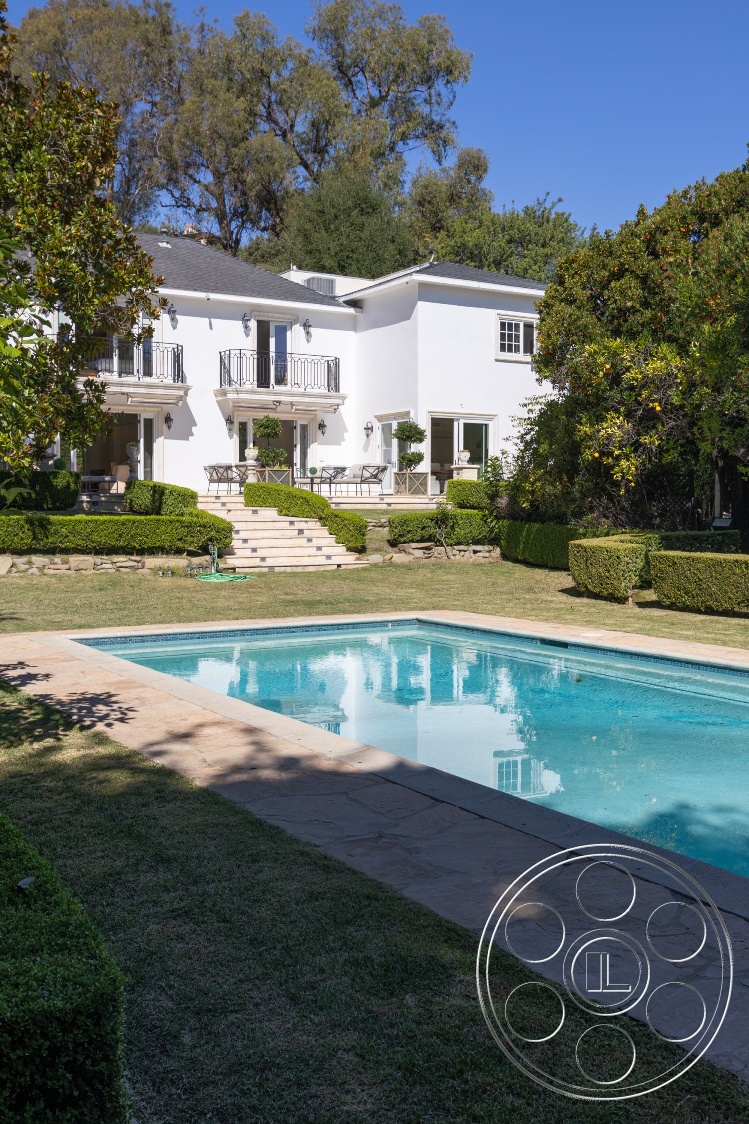Americana 431 - rectangular swimming pool, landscaped lawn, sunny outdoor space, balcony with wrought iron railings, stone pool deck, exterior white stucco walls, concrete pool border, tropical garden setting, planted garden with manicured hedges, terraced steps leading to double doors, exposed wooden deck seating area, lush green trees backdrop