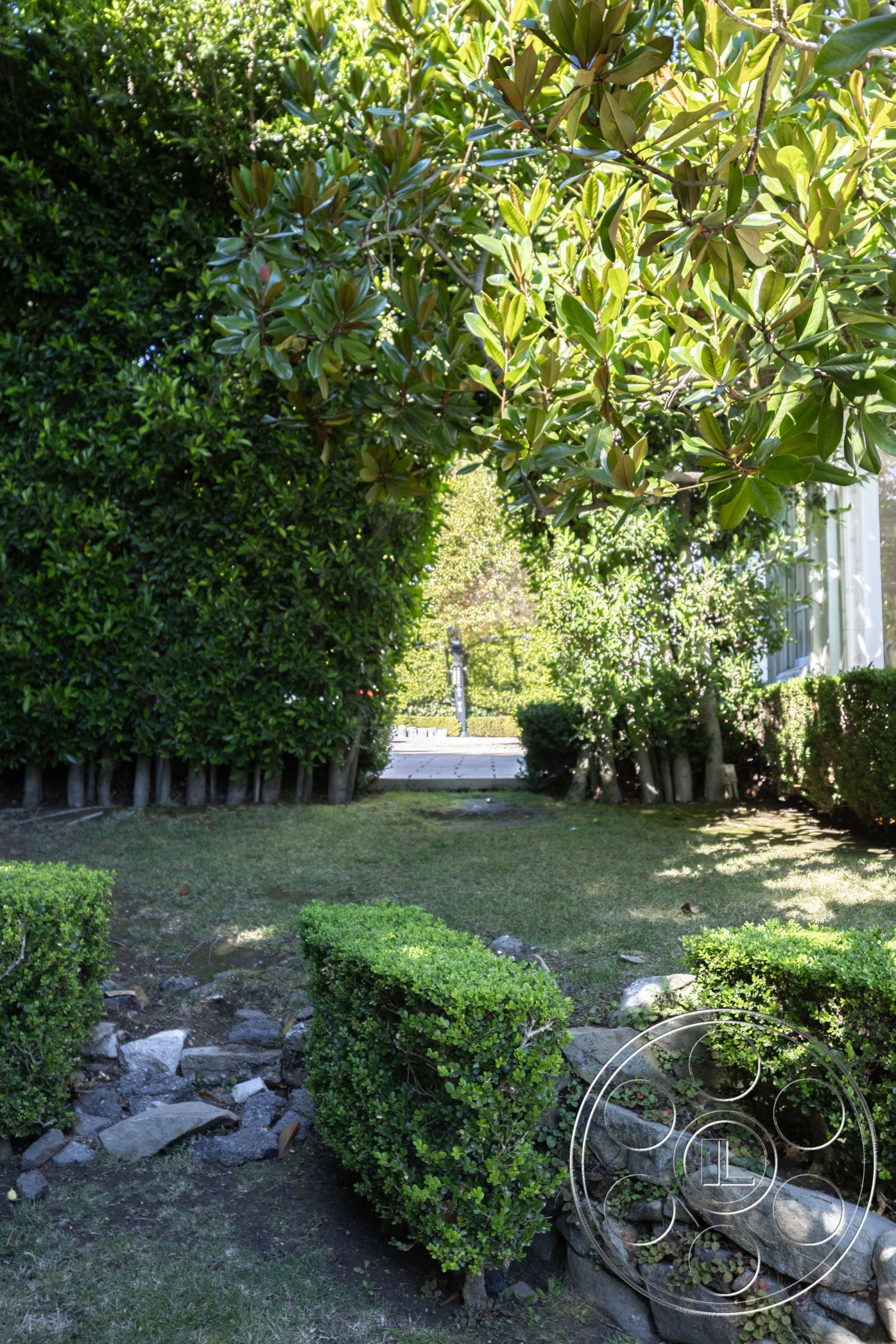 Americana 431 - surrounding foliage, paved stone walkway, exterior garden pathway, lush greenery setting, sunlit outdoor area, manicured grass lawn, hedge border landscaping, natural stone garden walls, bushes with rounded shapes, wooden fence posts, symmetrical landscaping design, outdoor green space, garden entryway, softscape with shrubs, textured stone path edges, decorative stone ground cover, garden retreat space