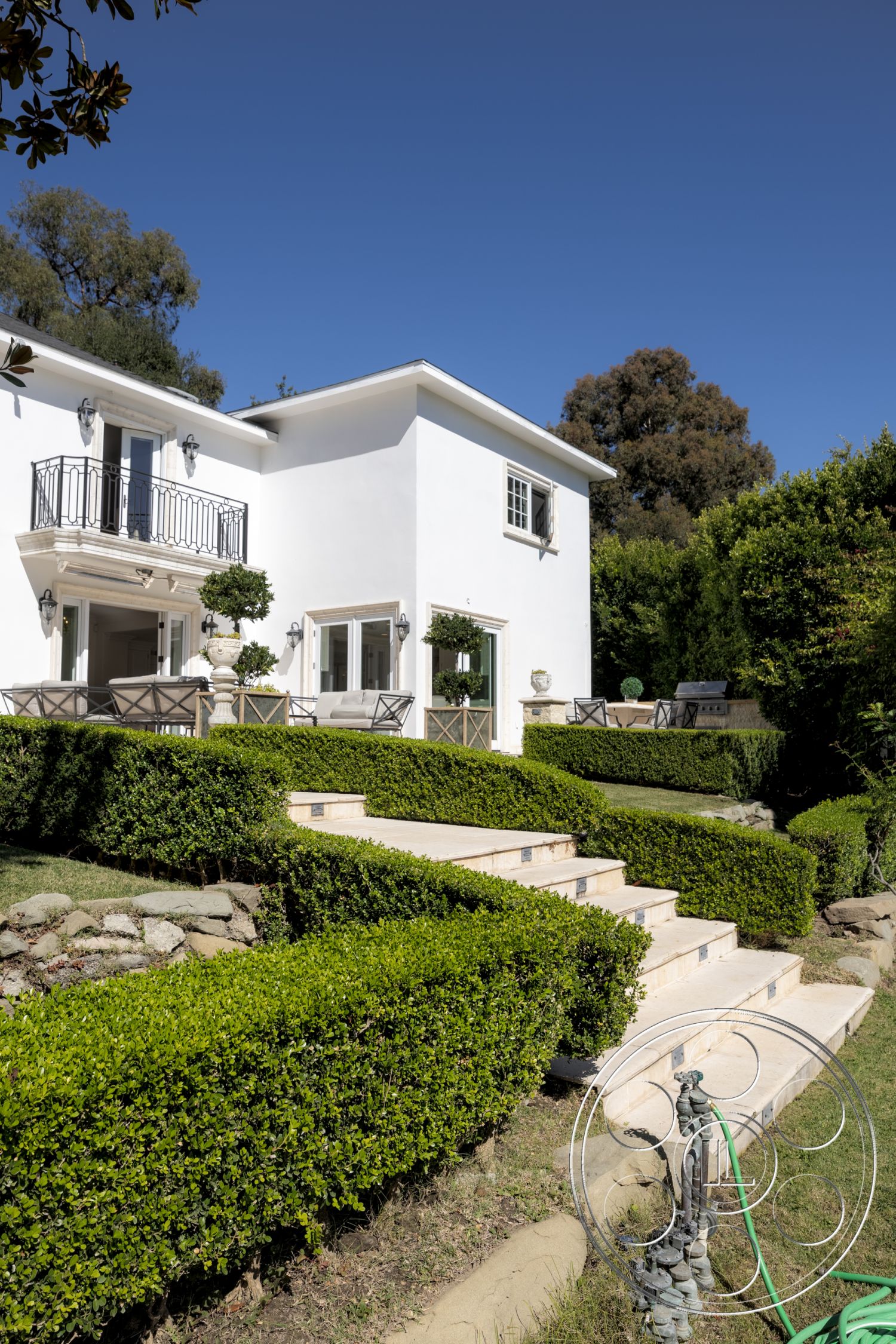 Americana 431 - outdoor seating area, floor-to-ceiling windows, urban residential setting, wooden outdoor furniture, decorative stone planter, modern Mediterranean style, landscaped garden setting, modern home architecture, green lawn area, black wrought iron balcony railing, natural stone wall accents, white stucco exterior walls, curved stair design, beige travertine steps, exposed aggregate stone pathway, manicured boxwood hedges, two-story home design, arched entryway details, sunny exterior patio