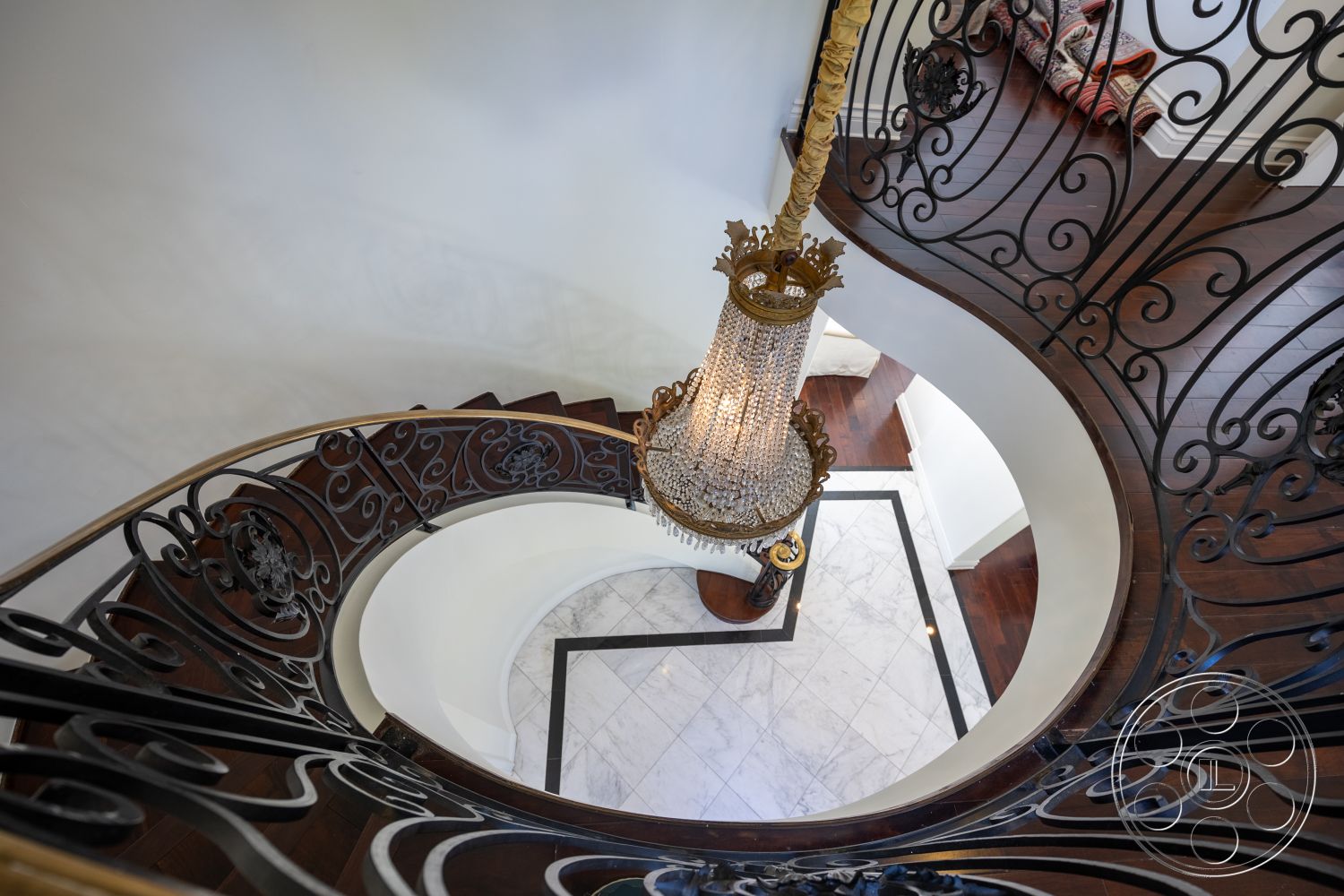 Americana 431 - high ceilings, open floor plan, textured wall finish, elegant lighting fixture, luxurious entryway, white painted walls, curved staircase design, interior spiral staircase, dark hardwood stair treads, wrought iron stair railings, chandelier with crystal details, marble flooring with black accent border