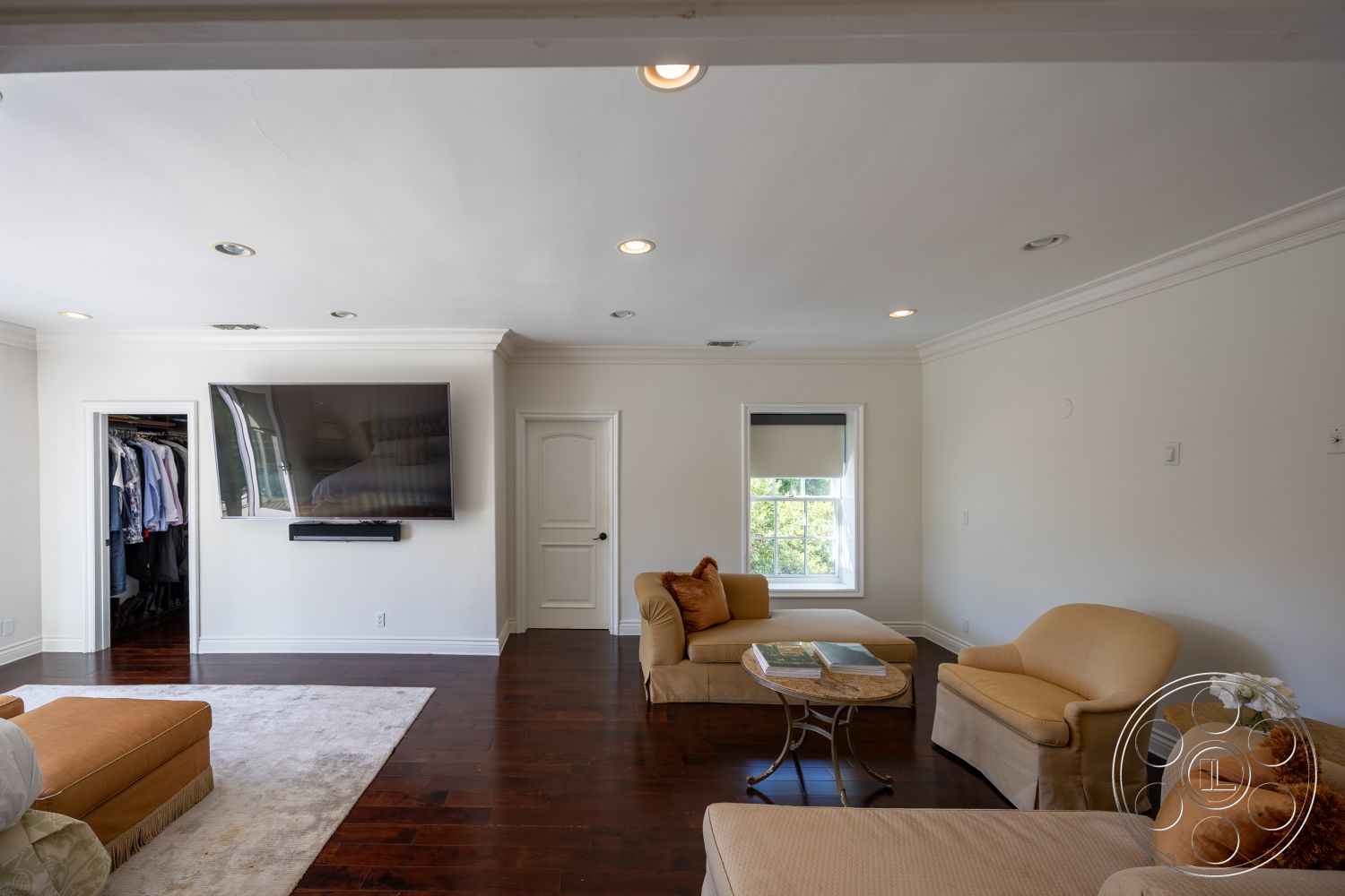 Americana 431 - hardwood bedroom floors, white painted walls, crown molding details, modern wall-mounted television, exposed ceiling detail, archway to closet space, double-hung window, beige upholstered seating, area rug for living room, functional home layout