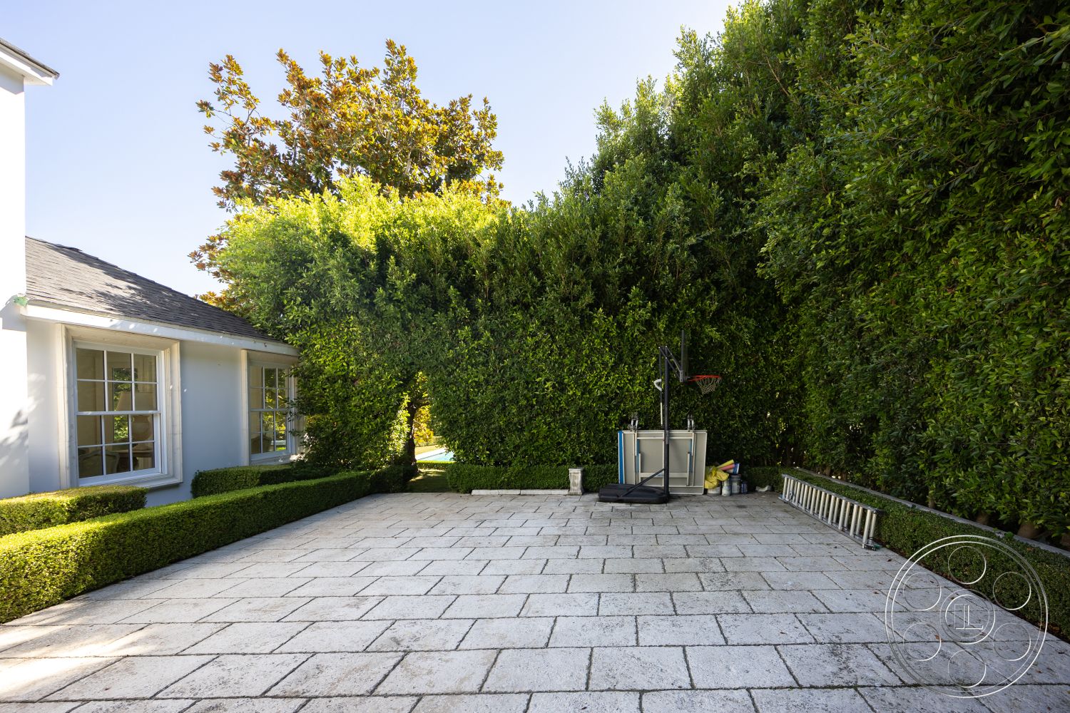 Americana 431 - minimalist aesthetic, open outdoor space, basketball area, urban backyard, hedge landscaping, flat roof design, white painted walls, rectangular patio layout, clean lines architecture, sunny outdoor environment, exterior patio flooring, boxwood hedge border, natural light entry, textured stone patio flooring, modern house exterior, lush green backdrop