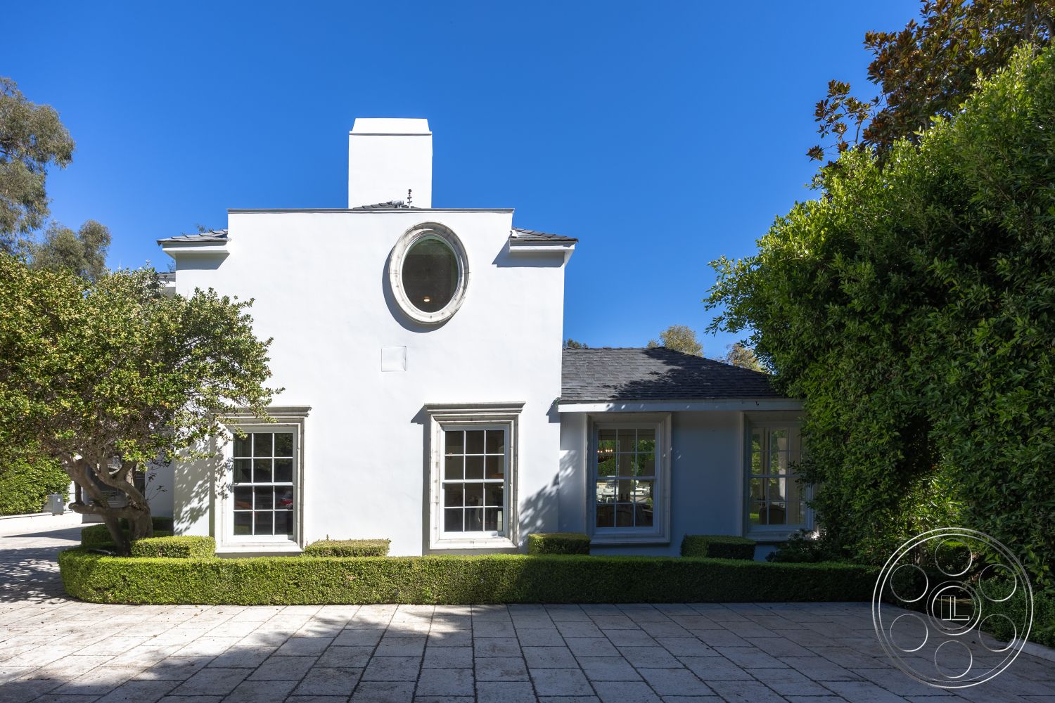 Americana 431 - exterior, white stucco walls, landscaped front yard, single-story home, symmetrical facade, oval window, boxwood hedges, slate roof, paved stone walkway, traditional window design, home with courtyard layout, modern classic architecture, city garden setting, front elevation with greenery