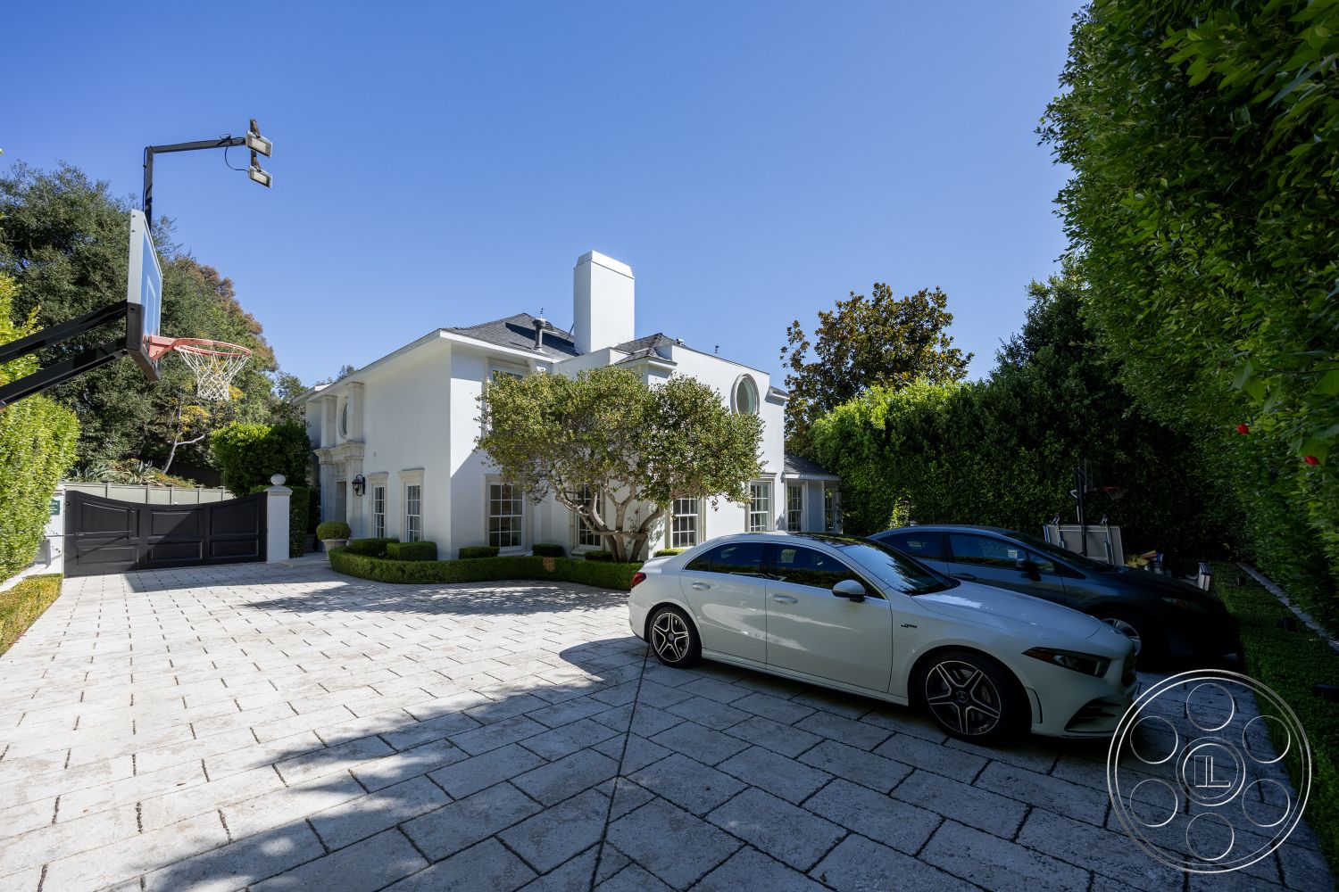 Americana 431 - exterior, white stucco walls, contemporary home, arched windows with decorative trim, lush greenery surrounding the property, modern residential design, paved driveway with large stone pavers, two-story home with a hip roof, basketball hoop on black steel post, landscaped front yard with rounded hedges, courtyard area with smooth stone paving, black wooden gate at driveway entrance, light-colored window frames