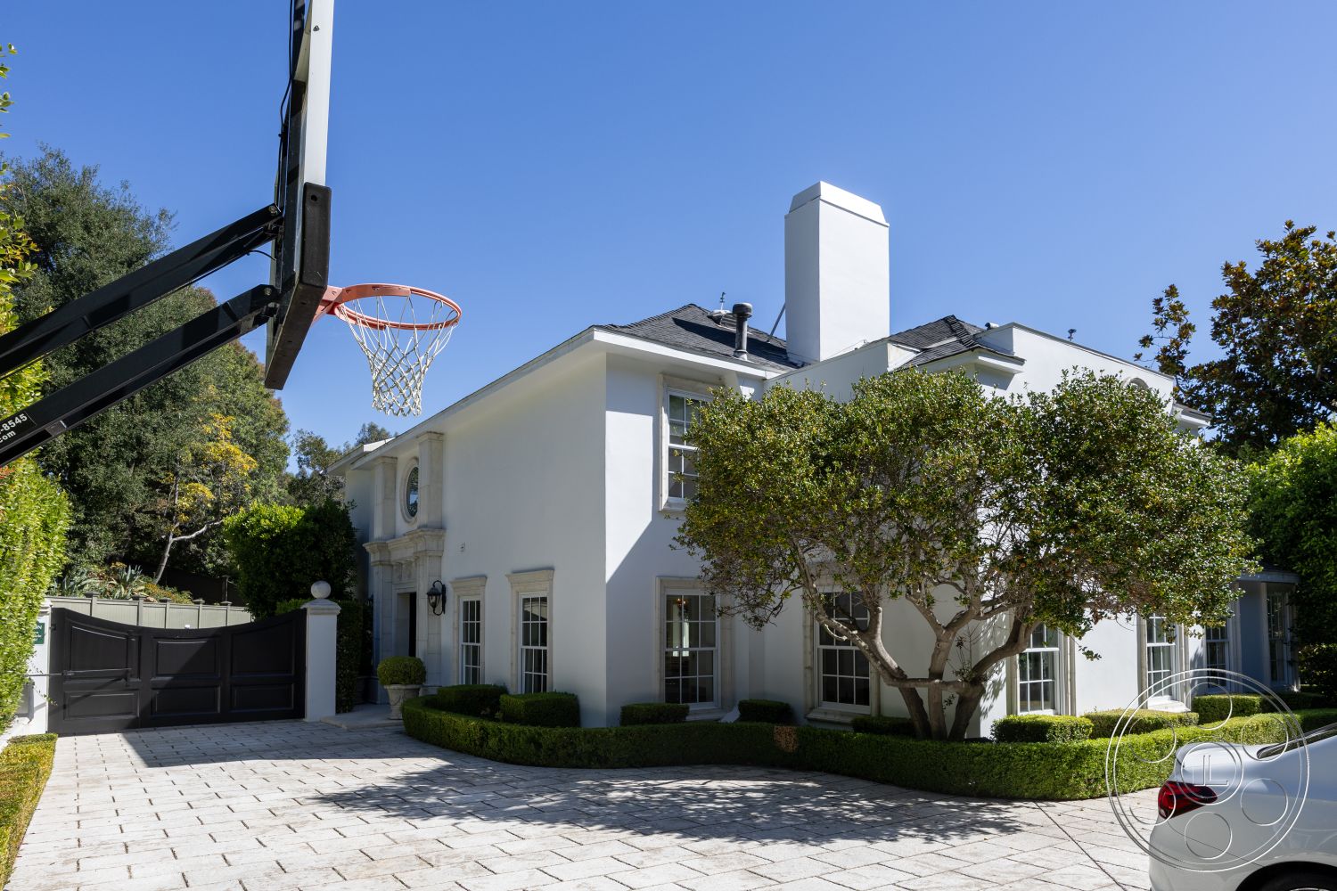 Americana 431 - suburban setting, gray slate roof, cobblestone driveway, exterior white stucco walls, arched windows with decorative trim, sunny outdoor environment, symmetrical facade design, rectangular driveway layout, landscaped front yard with hedges, ornamental tree in front yard, black metal driveway gate, classic lantern wall sconces, brick chimney stack, modern basketball hoop installation, paved stone walkways, detailed molding around windows, lush green shrubbery landscaping, two-story home structure
