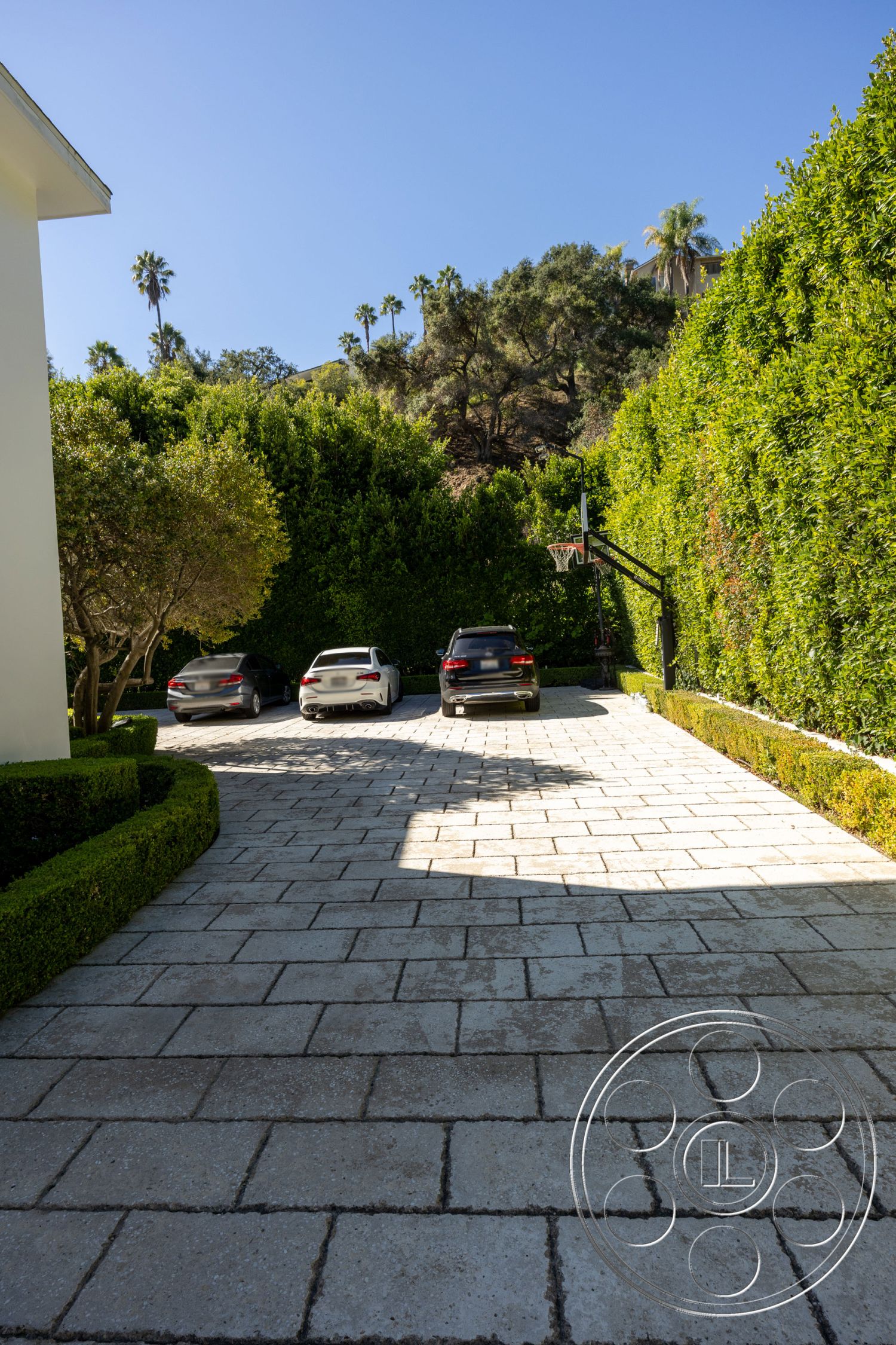 Americana 431 - exterior, paved driveway, surrounding greenery, suburban setting, curved driveway, surrounding palm trees, landscaped hedges, textured concrete pavers, basketball hoop installation, central circular design in driveway, sunlit landscape, neatly trimmed boxwood shrubs, elevated hillside, modern residential driveway