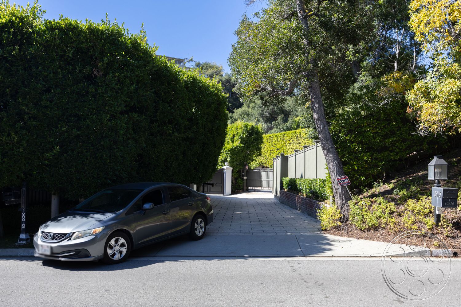 Americana 431 - suburban neighborhood, landscaped front yard, privacy landscaping, outdoor lighting fixtures, urban residential, concrete sidewalk, curved driveway design, tropical foliage landscaping, exterior driveway, paved driveway stones, green hedges fence, brick planter bed, wood fence entrance, single-family home setting, adjoining tree cover, light traffic street