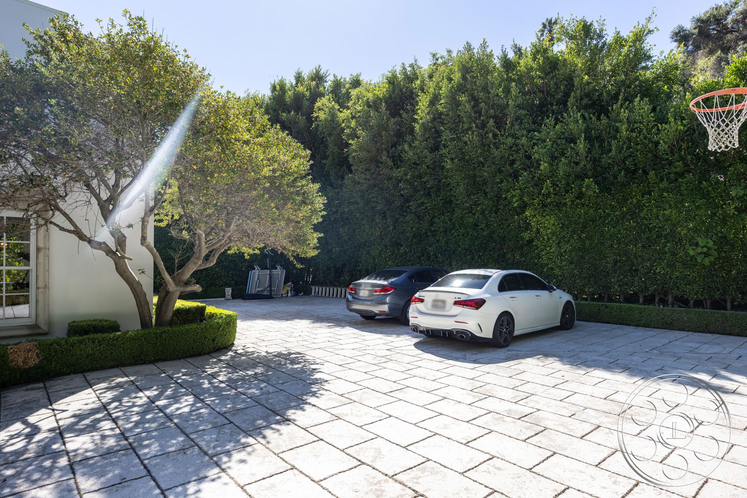 Americana 431 - exterior, smooth stone surface, lush greenery backdrop, landscaped hedges, sunlit outdoor space, surrounding urban landscape, modern home entrance, basketball hoop installation, stamped concrete patio floor, stone paver driveway, rectangular driveway design, mature trees shading driveway, two parked vehicles