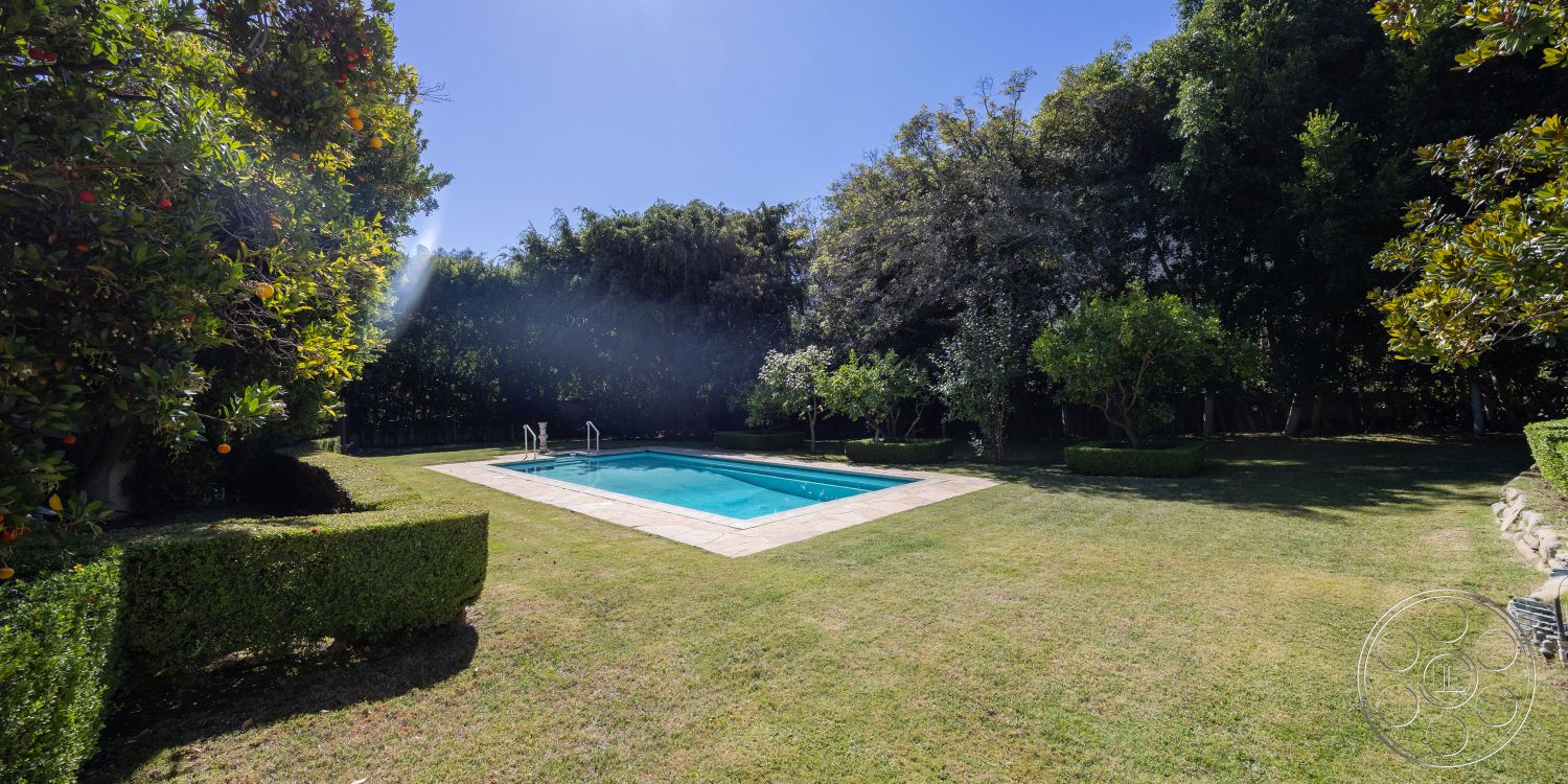 Americana 431 - exterior, landscaped garden, lush green lawn, manicured hedges, outdoor relaxation area, rectangular pool, concrete pool border, sunlit outdoor space, concrete patio surround, dense tree coverage, modern swimming pool design, family-friendly backyard