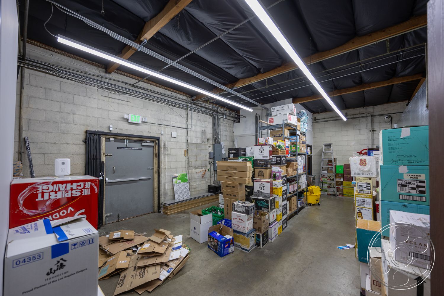 Shop 127 - open layout, concrete flooring, functional storage, utility area, interior storage space, stacked cardboard boxes, exposed concrete block walls, industrial shelving units, warehouse setting, metal beam ceiling, vinyl strip lighting, fire exit door, cluttered organization, modern utility space