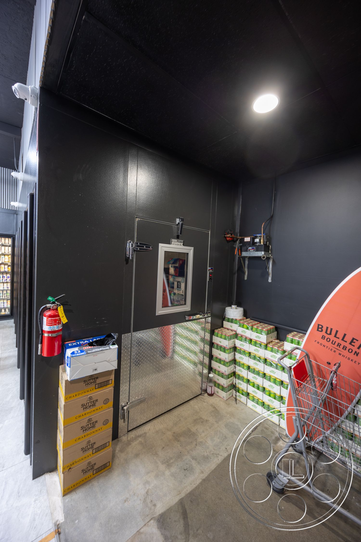 Shop 127 - industrial design elements, black painted walls, organized storage space, fluorescent ceiling lighting, fire extinguisher mounted on wall, concrete floor with polished finish, interior storage room, metal door with window, diamond plate door surface, shelves stacked with boxes, cart for transporting goods, cooler access door, adjacent room entrance, warehouse-like atmosphere, functional retail area